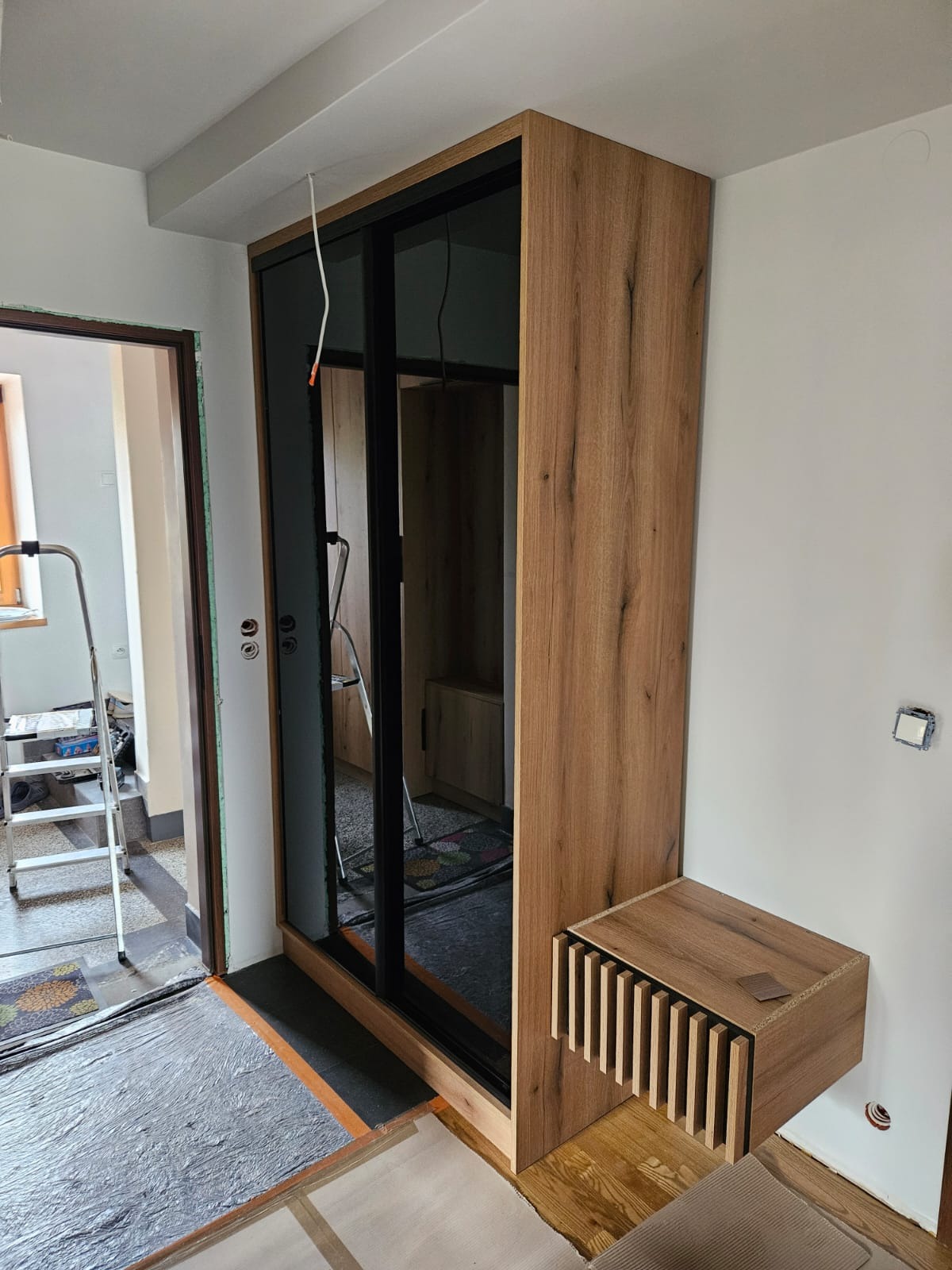 Montaż szafy przesuwnej z drewnianym dekorem i czarnymi ramami. Widoczna szafka nocna z lamelami. W tle drabina i fragment remontowanego pomieszczenia.