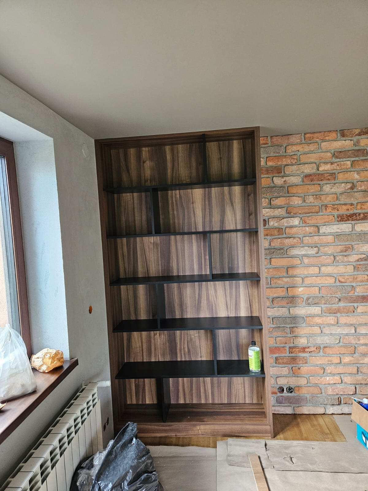 Wysoka, drewniana szafa wnękowa z czarnymi półkami, w surowym wnętrzu z ceglaną ścianą. Widoczne niedokończone wykończenia i materiały budowlane.
