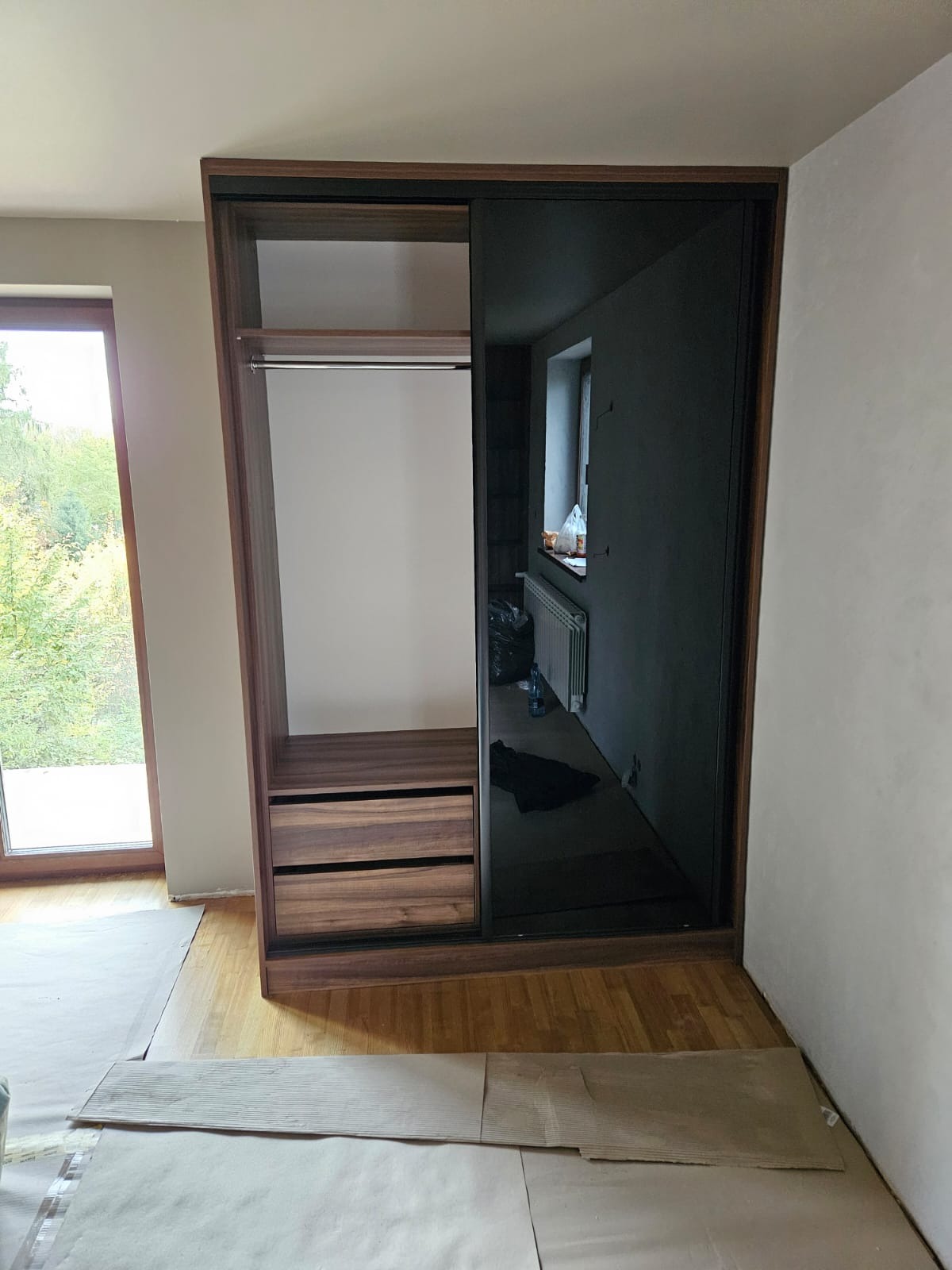 Wnętrze z szafą wnękową z przesuwnymi drzwiami, wykończoną drewnem i czarnym szkłem. Widoczne półki, szuflady i drążek na ubrania. Podłoga zabezpieczona kartonem.