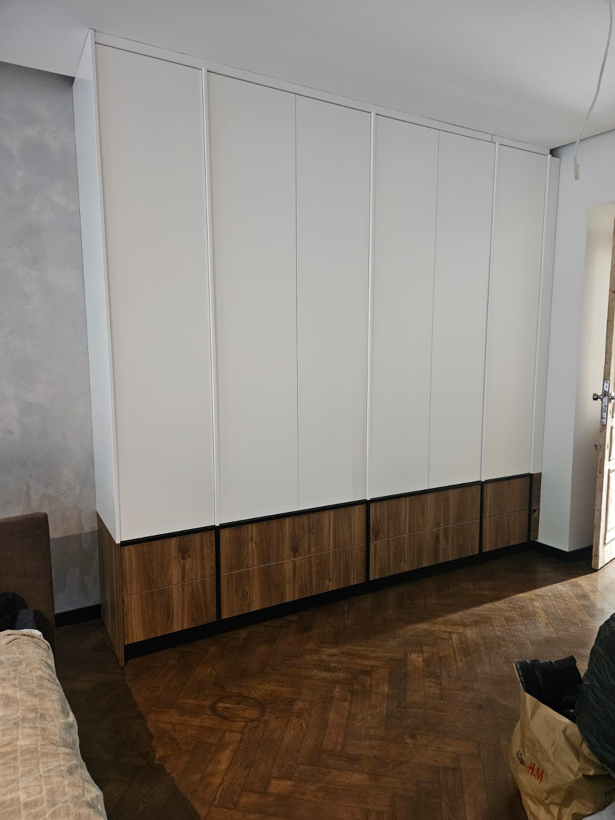 Biała szafa wnękowa z drewnianymi frontami dolnymi, widoczna na tle parkietu i fragmentu pokoju. Minimalistyczny design, proste linie, naturalne materiały.