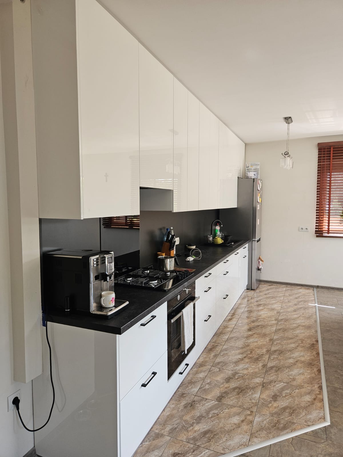 Biała kuchnia z połyskiem, czarny blat, nowoczesne uchwyty. Ekspres do kawy na blacie. Widok na okno z drewnianymi żaluzjami. Minimalistyczny design.