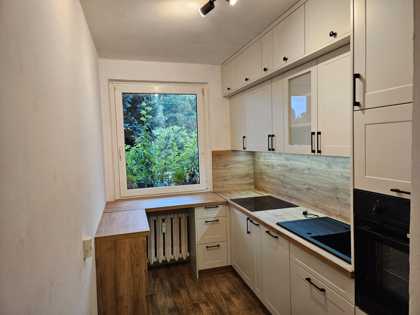 Jasna kuchnia na wymiar z drewnianym blatem i białymi szafkami, z widokiem na zieleń za oknem. Grzejnik wkomponowany w zabudowę. Minimalistyczne uchwyty.