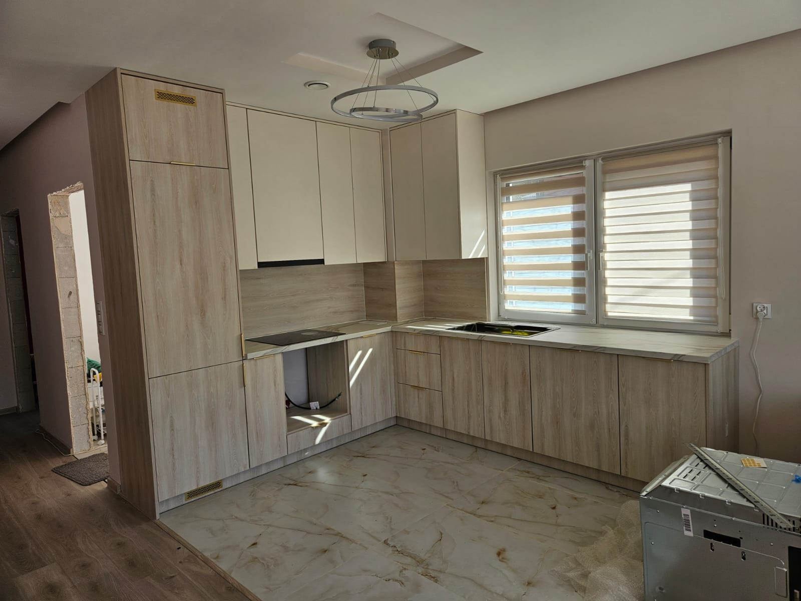 Jasna kuchnia z drewnopodobnymi frontami i marmurowym blatem, oświetlona nowoczesną lampą. Widoczne okno z żaluzjami i fragment niezakończonego montażu sprzętu AGD.