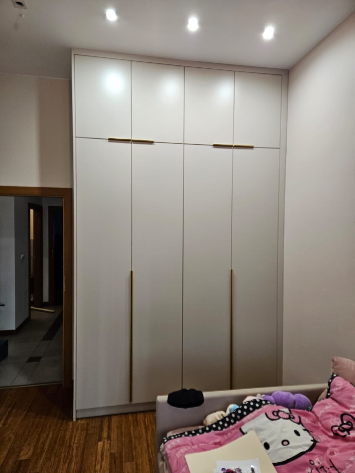 Jasna szafa wnękowa z długimi, złotymi uchwytami, widoczna w sypialni w Kalwarii Zebrzydowskiej. Minimalistyczny design, drewniana podłoga, drzwi do innego pokoju w tle.