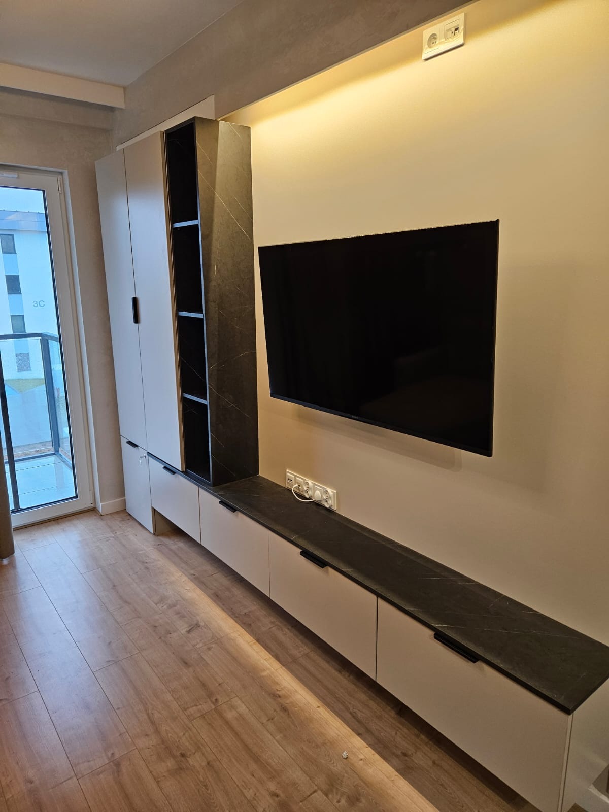 Nowoczesna zabudowa meblowa z szafkami i półkami w salonie, wykończona w bieli i czarnym marmurze, z telewizorem na ścianie i drewnianą podłogą.