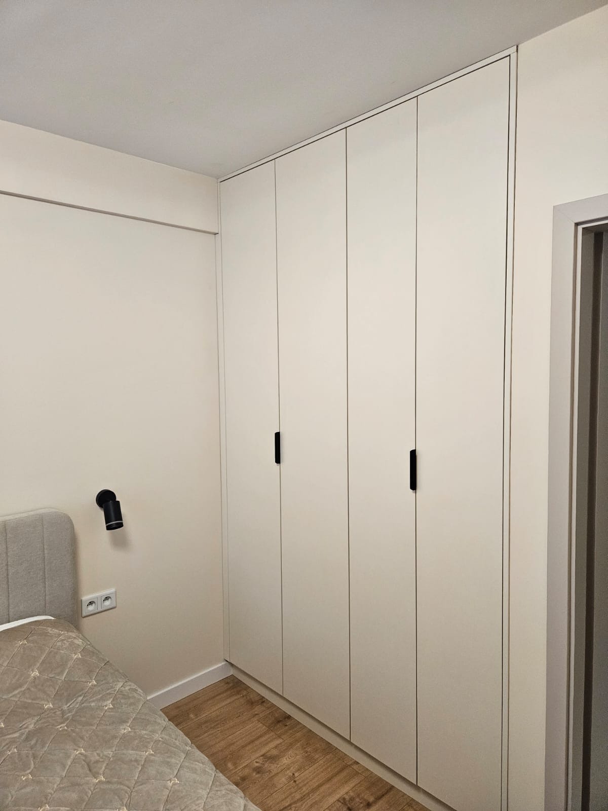 Biała szafa wnękowa z czterema drzwiami i czarnymi uchwytami, dopasowana do ściany w sypialni. Widoczny fragment łóżka i lampka nocna.