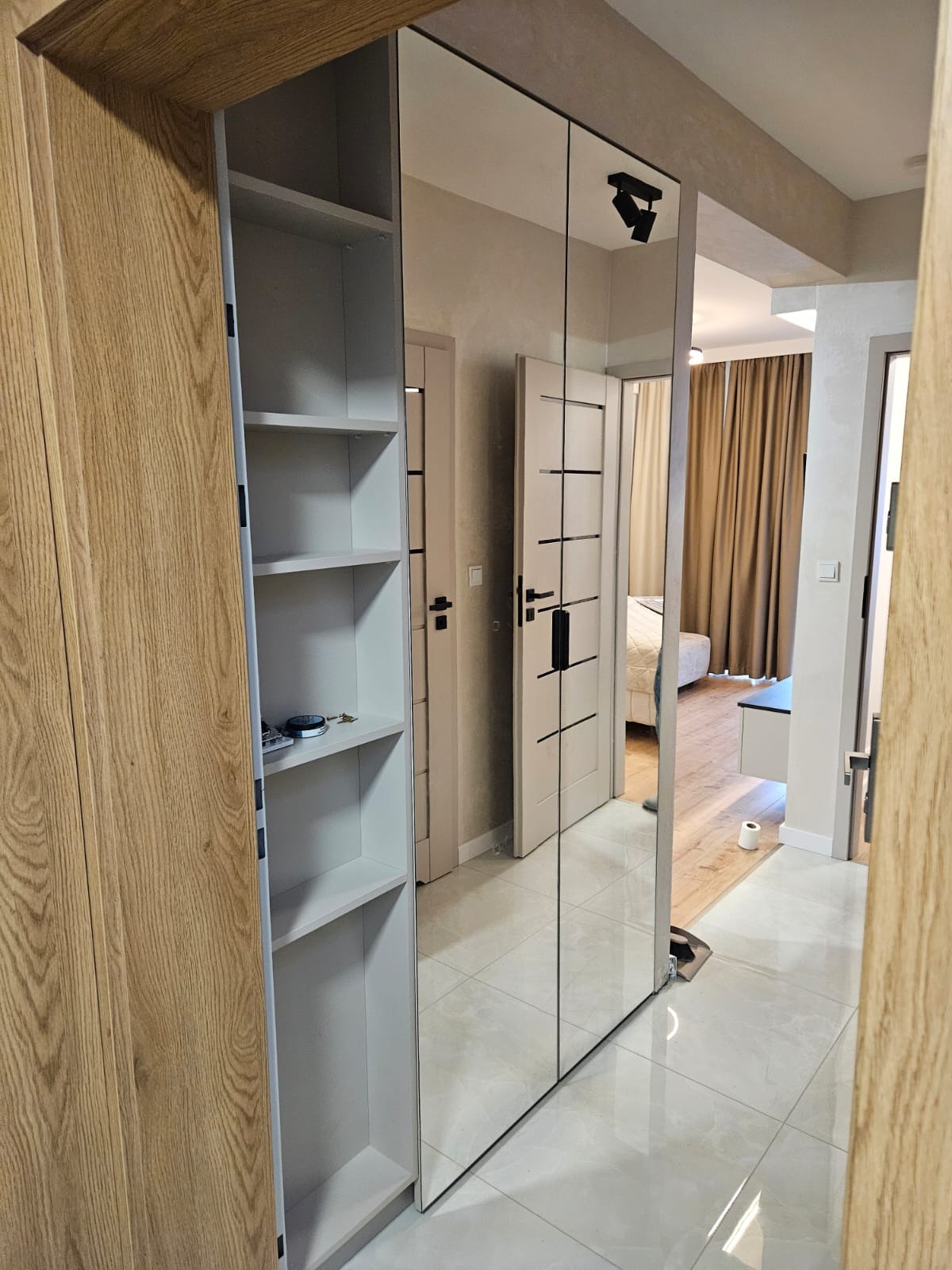 Nowoczesna szafa wnękowa z lustrzanymi drzwiami i otwartym regałem z jasnego drewna, odbijająca wnętrze pokoju z łóżkiem i zasłonami.