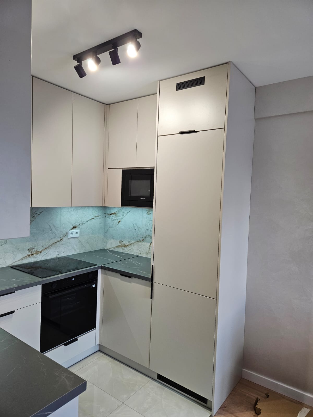 Jasna kuchnia z szarym blatem i marmurowym backsplashiem. Czarna płyta indukcyjna i piekarnik. Oświetlenie punktowe na suficie. Minimalistyczne uchwyty.