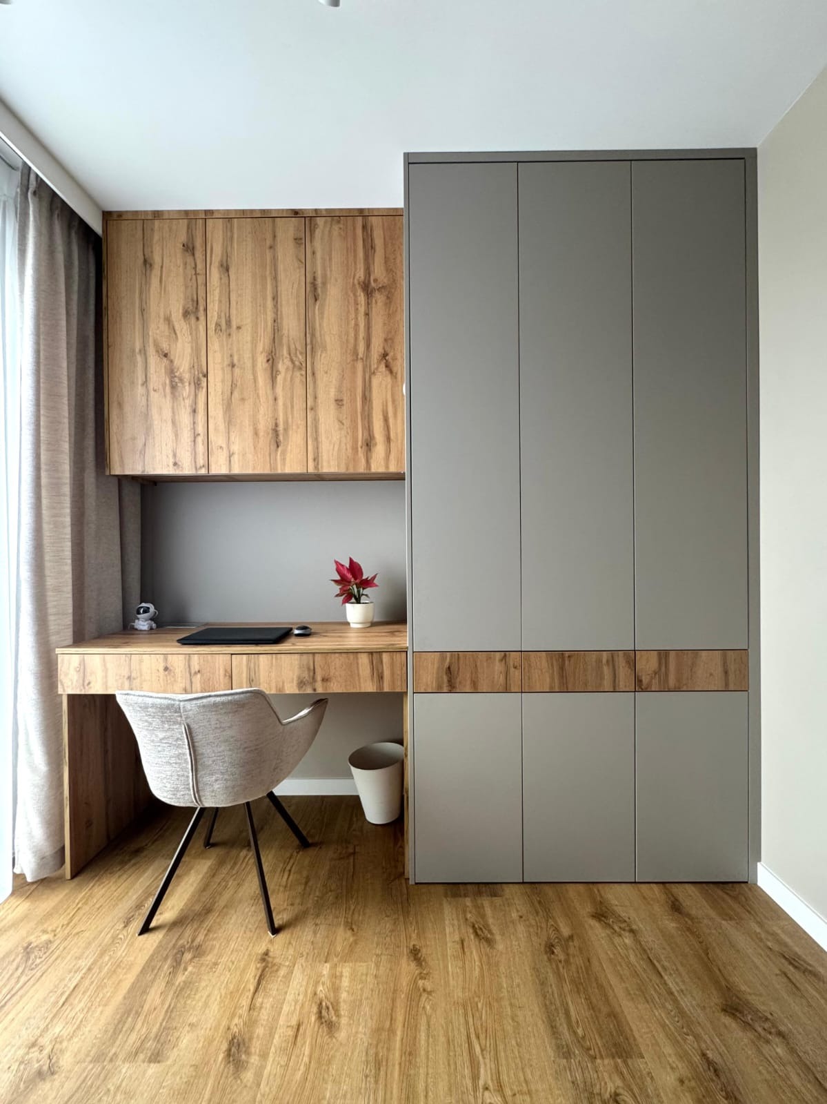 Nowoczesny pokój z biurkiem z drewnianym blatem i szarą szafą w zabudowie. Na biurku laptop, roślina w doniczce i figurka. Jasna podłoga i krzesło.
