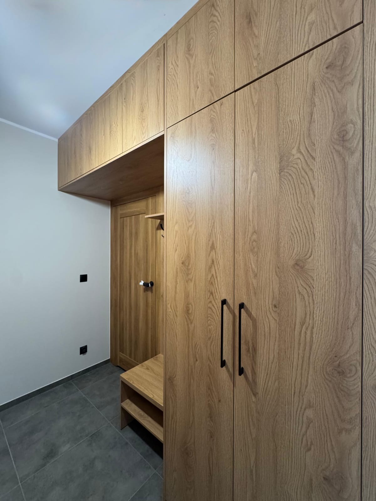 Drewniana szafa wnękowa z siedziskiem i pawlaczem, czarne uchwyty, widok na drzwi. Realizacja w przedpokoju, szara podłoga.