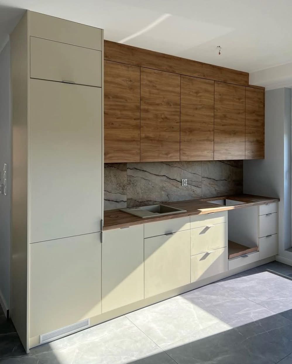 Nowoczesna kuchnia z beżowymi szafkami dolnymi i drewnopodobnymi górnymi, marmurowy backsplash, szare płytki podłogowe i subtelne oświetlenie.