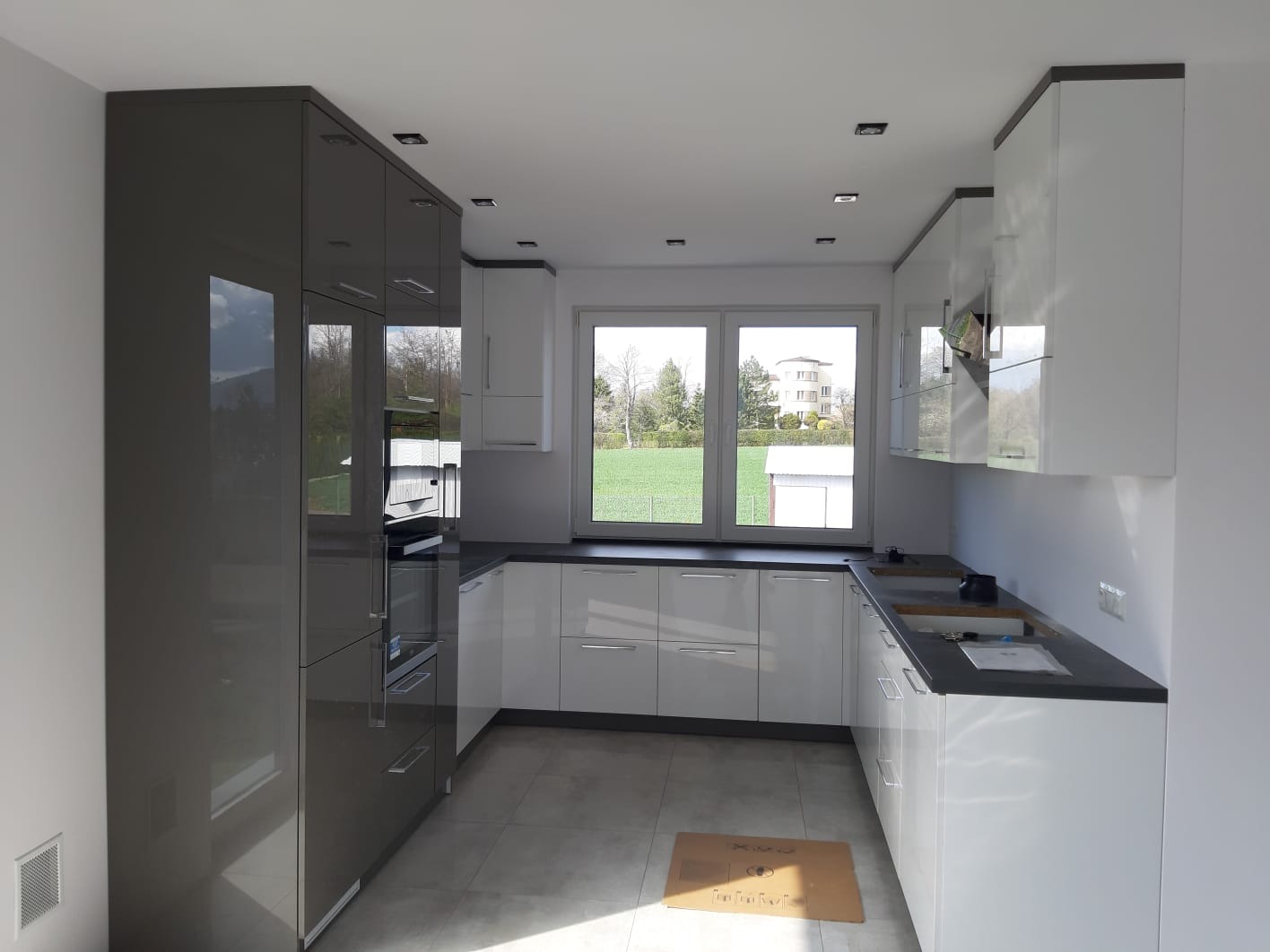 Nowoczesna kuchnia z połyskiem: szare wysokie szafki i białe dolne, czarny blat, duże okno z widokiem na zieleń. Minimalistyczny design, jasne wnętrze.