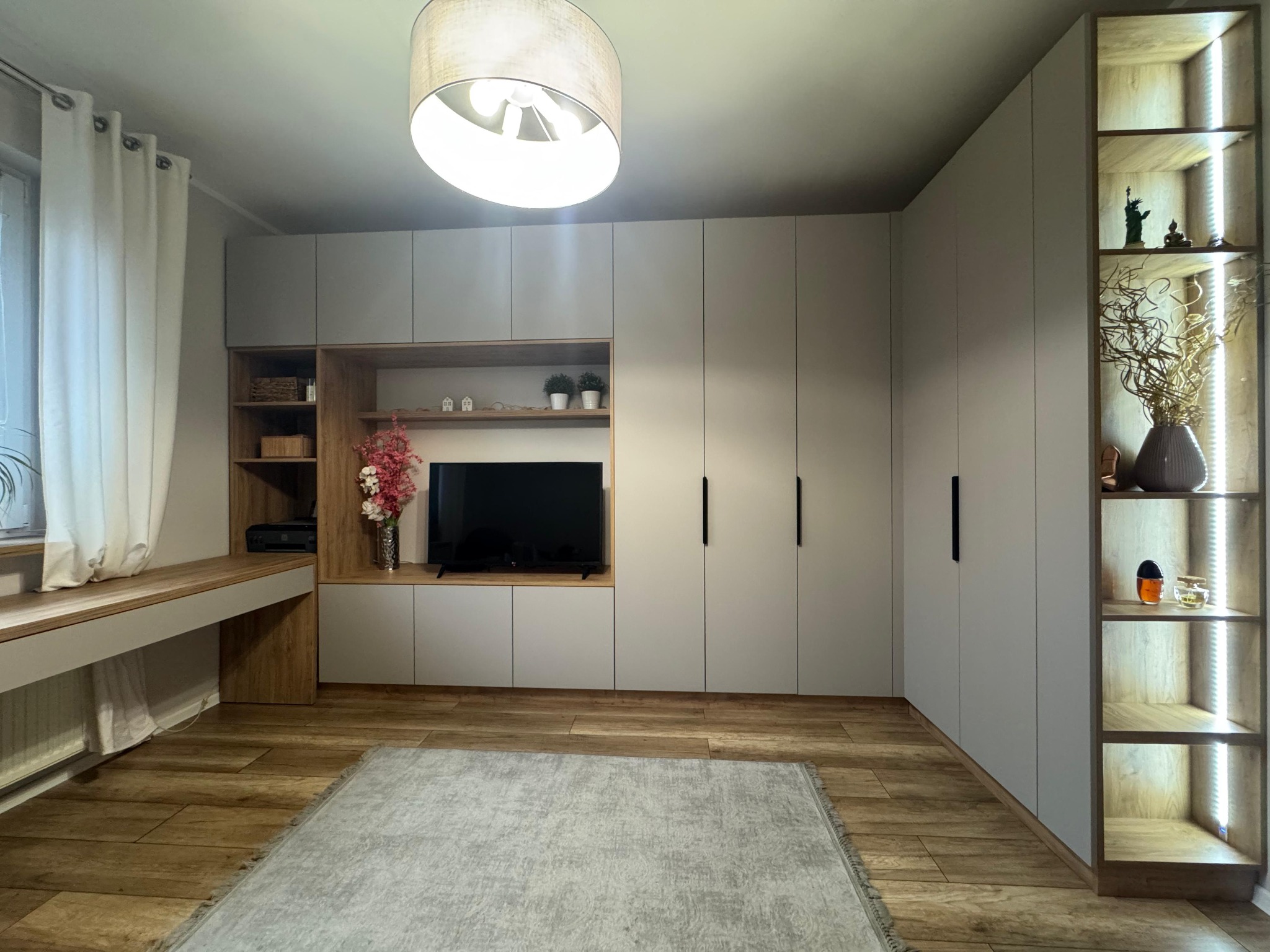 Jasnoszara zabudowa meblowa z szafami, półkami i biurkiem, oświetlona lampą sufitową. Drewniana podłoga i dywan w odcieniach szarości. Minimalistyczny design.