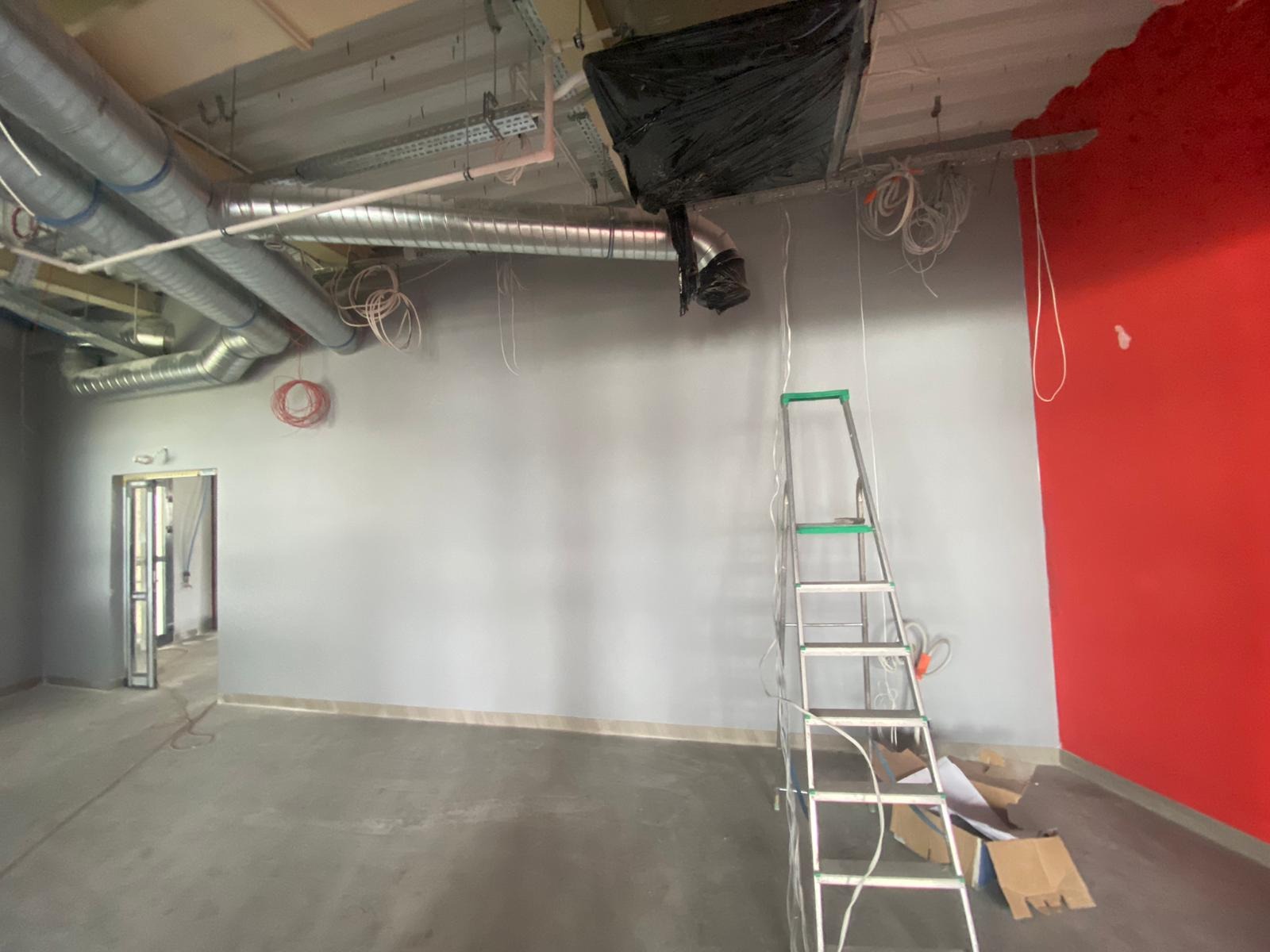 Wnętrze w trakcie prac wykończeniowych: szare ściany, czerwona ściana akcentująca, instalacje pod sufitem, drabina i kartony na podłodze. Widok z niskiego kąta.