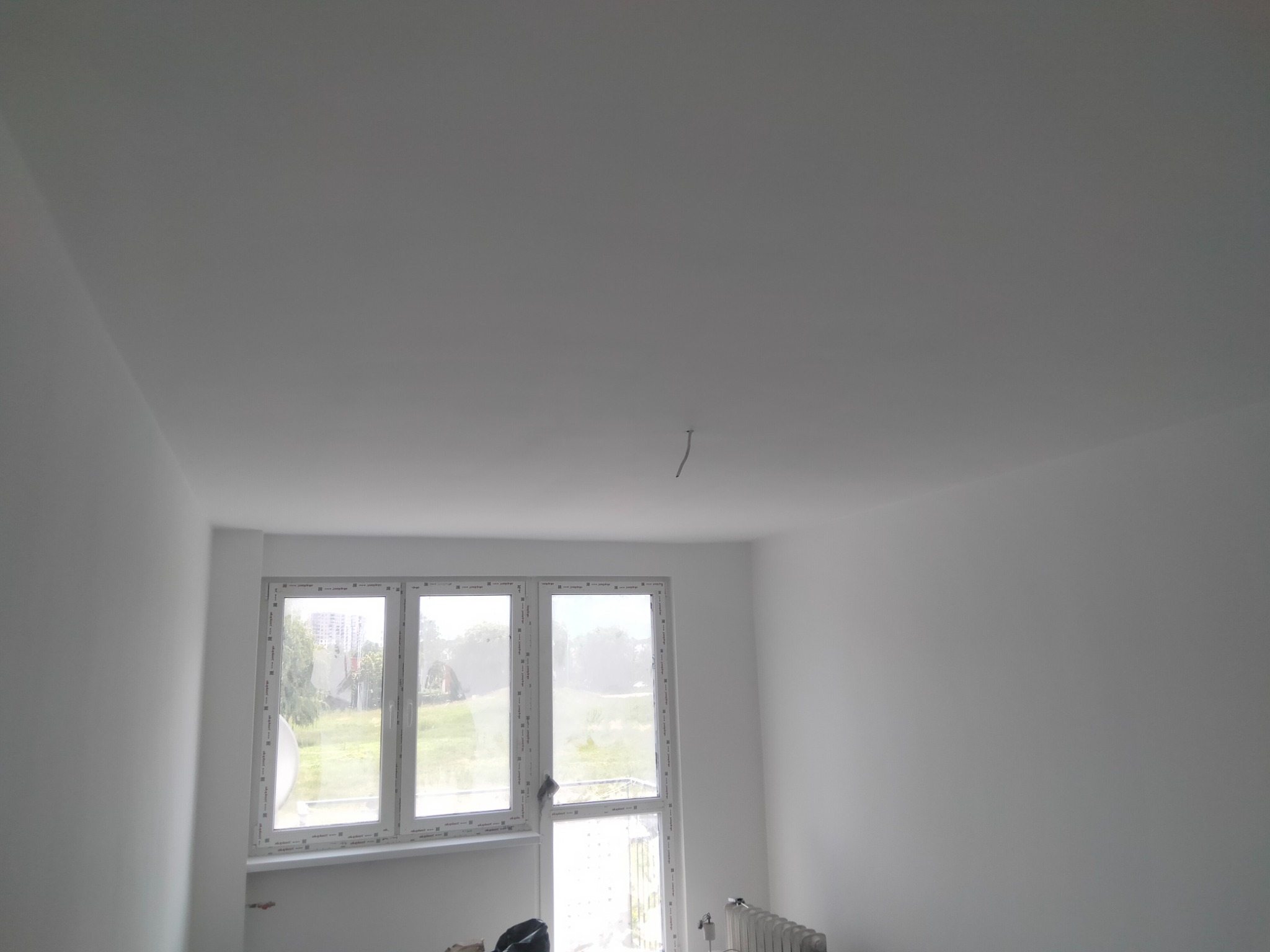 Wnętrze po malowaniu: białe ściany i sufit, okno z widokiem na zieleń. Minimalistyczny design, świeżo odnowione pomieszczenie z grzejnikiem i elementami malarskimi.