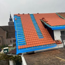 APPROVED GROUP SPÓŁKA Z OGRANICZONĄ ODPOWIEDZIALNOŚCIĄ - Częściowy widok na dach z czerwoną dachówką w trakcie wymiany, z widoczną membraną w kolorze niebieskim i łatami, w tle wieża kościelna.