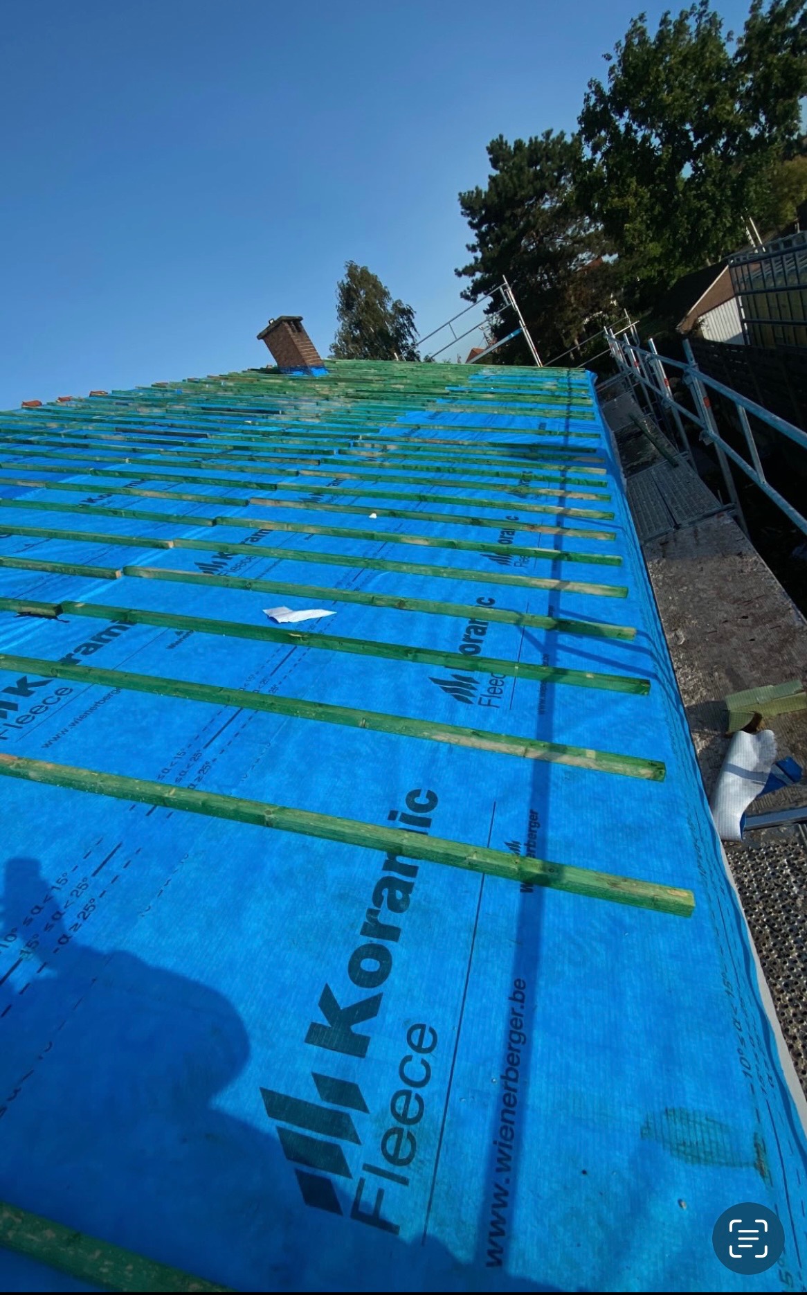 Dach w trakcie krycia, widoczna niebieska membrana Koramic Fleece i zielone łaty, komin w tle, ujęcie z dołu pod kątem.