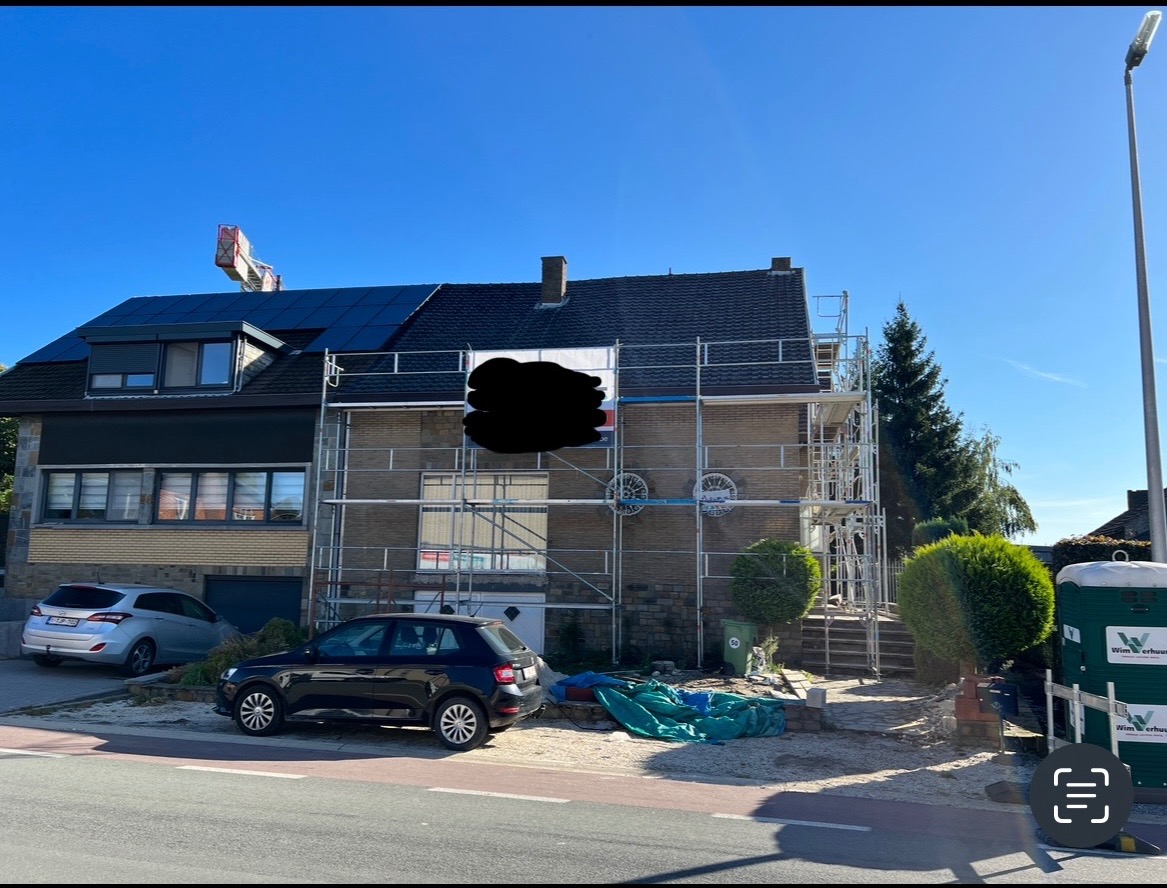 Dom jednorodzinny w trakcie remontu dachu, z widocznymi rusztowaniami i nowymi panelami słonecznymi na części dachu, na tle błękitnego nieba.