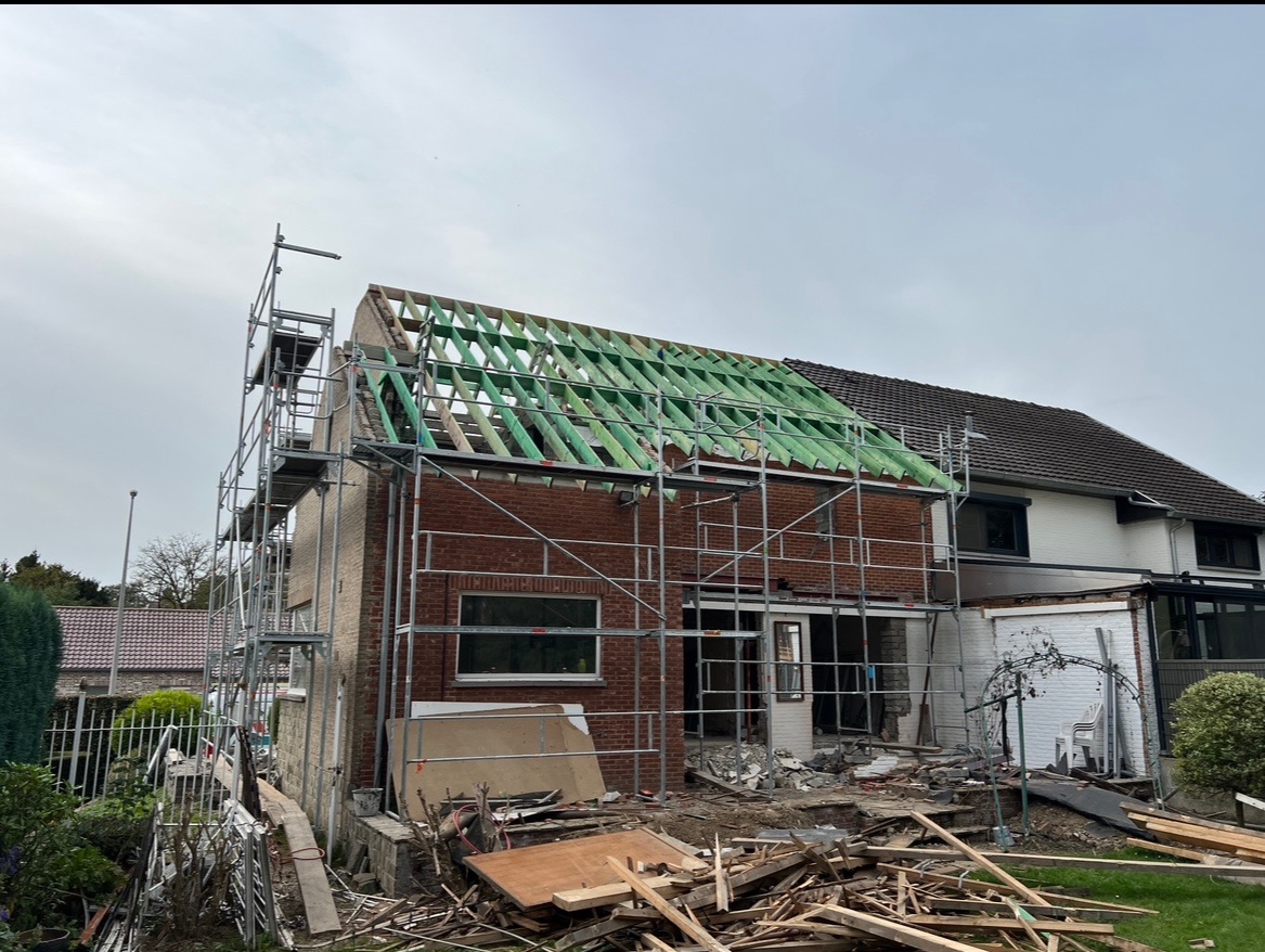 Budowa dachu z zielonymi elementami konstrukcyjnymi na ceglanym budynku, widoczne rusztowania i bałagan na placu budowy.