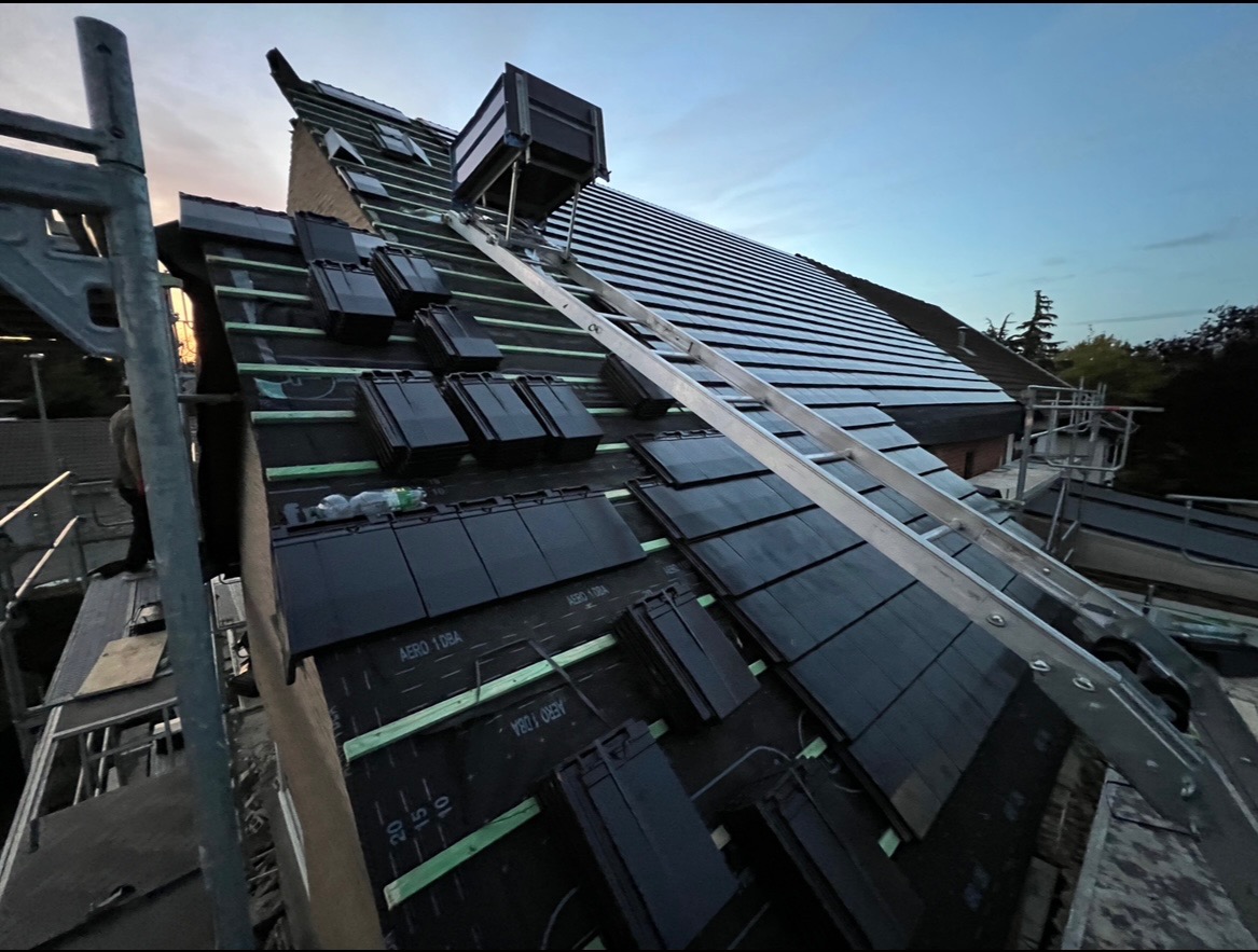 Montaż czarnej dachówki na dachu domu jednorodzinnego, widoczne rusztowanie i drabina, częściowo ułożone pokrycie dachowe z widoczną membraną dachową i łatami.