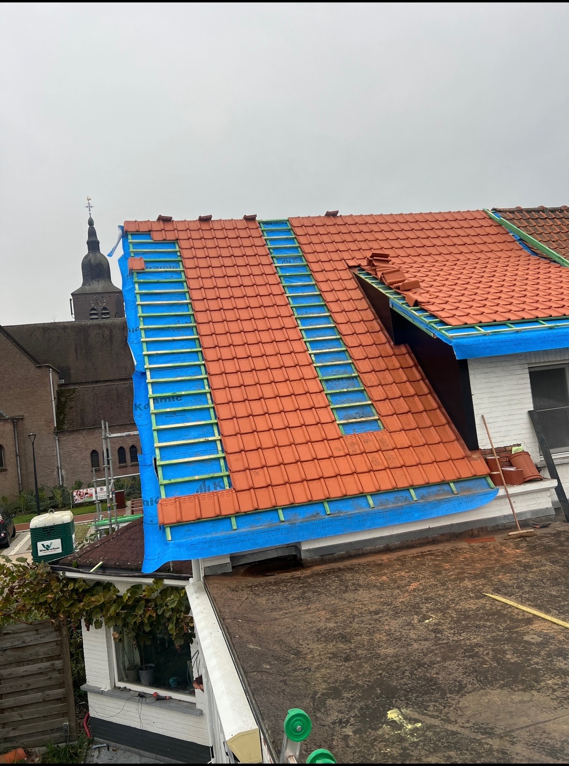Częściowy widok na dach z czerwoną dachówką w trakcie wymiany, z widoczną membraną w kolorze niebieskim i łatami, w tle wieża kościelna.
