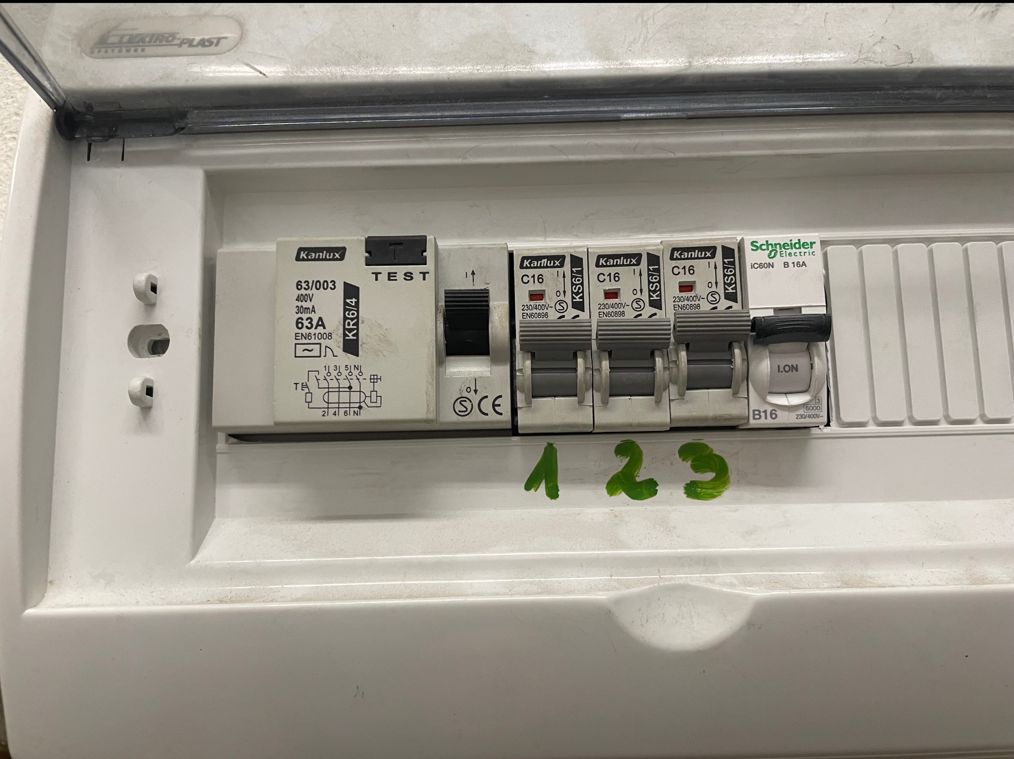 Rozdzielnica elektryczna z bezpiecznikami Kanlux i Schneider Electric, z zielonymi numerami '1 2 3' naniesionymi na obudowę. Widoczne oznaczenia parametrów technicznych.