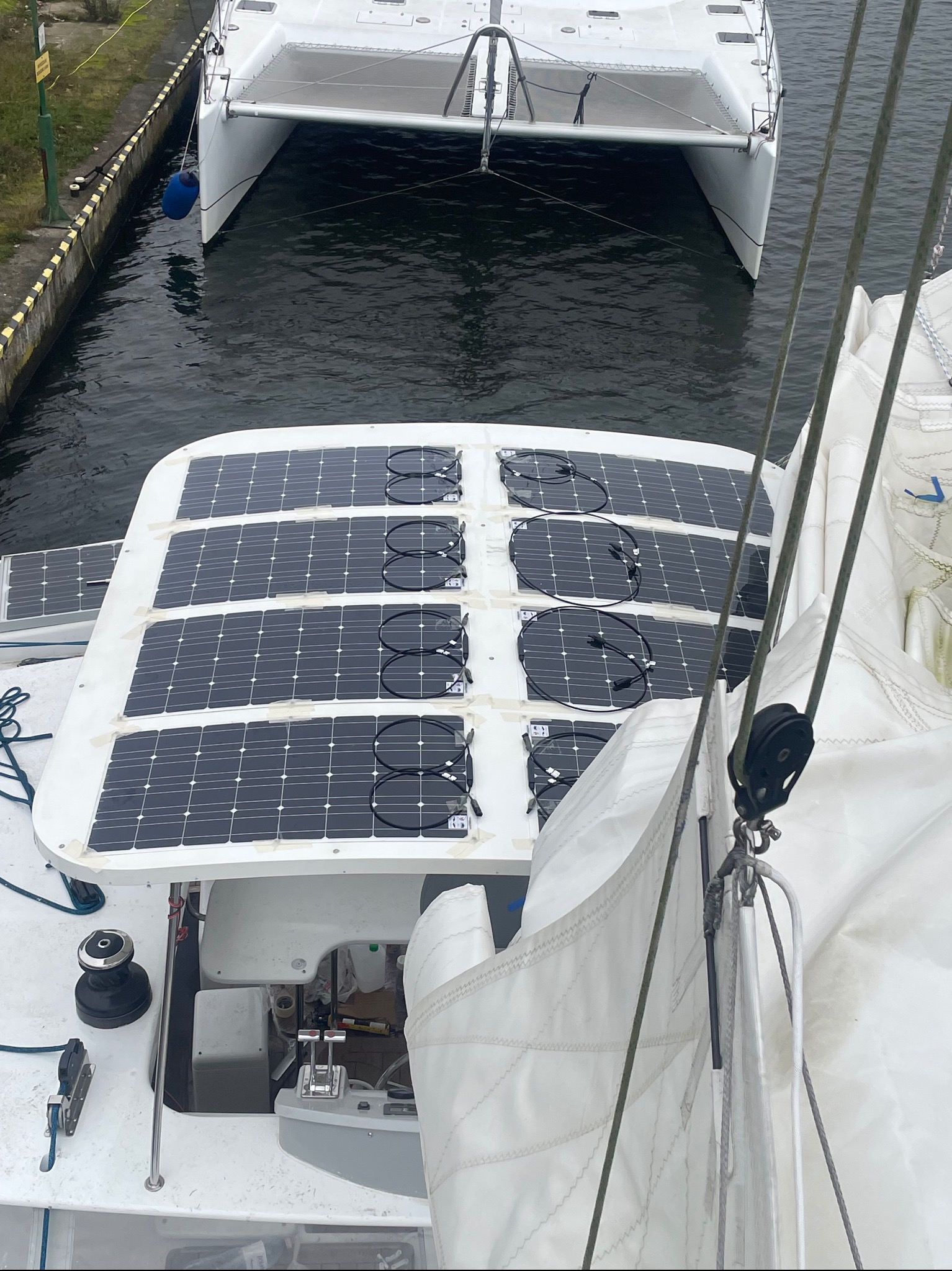 Widok z góry na panele słoneczne zamontowane na dachu jachtu, z widocznymi kablami i elementami mocującymi. W tle drugi jacht na wodzie.