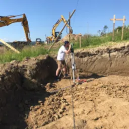 Pracownik w białej koszulce i beżowych szortach obsługuje niwelator na statywie, w tle wykop i dwie koparki JCB na placu budowy.