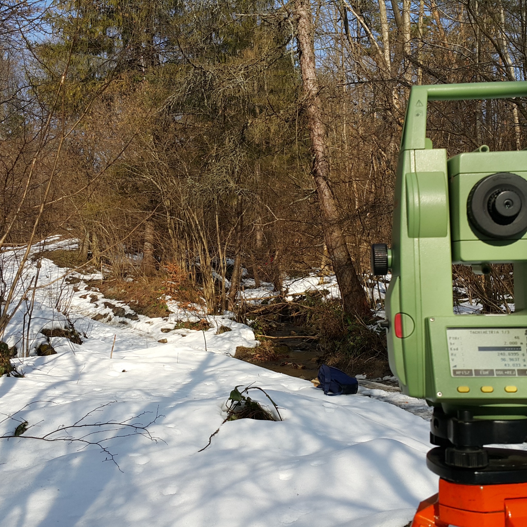 Zielony tachimetr na statywie, ustawiony w zasnieżonym terenie leśnym, z widocznym strumieniem w tle.