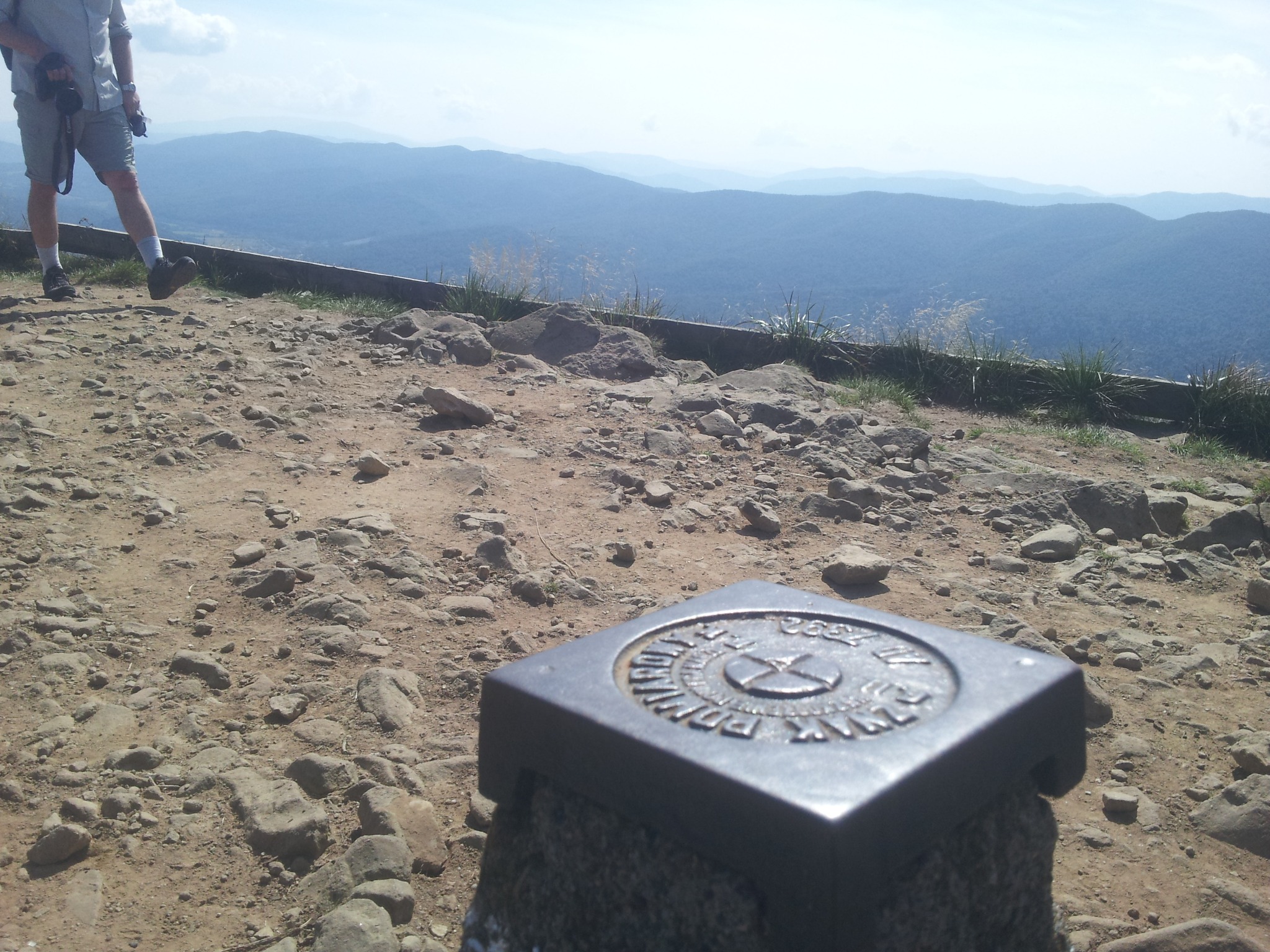 Metalowy słupek geodezyjny z wygrawerowanymi oznaczeniami, na szczycie góry z widokiem na zalesione wzgórza w oddali, częściowo widoczna postać w tle.