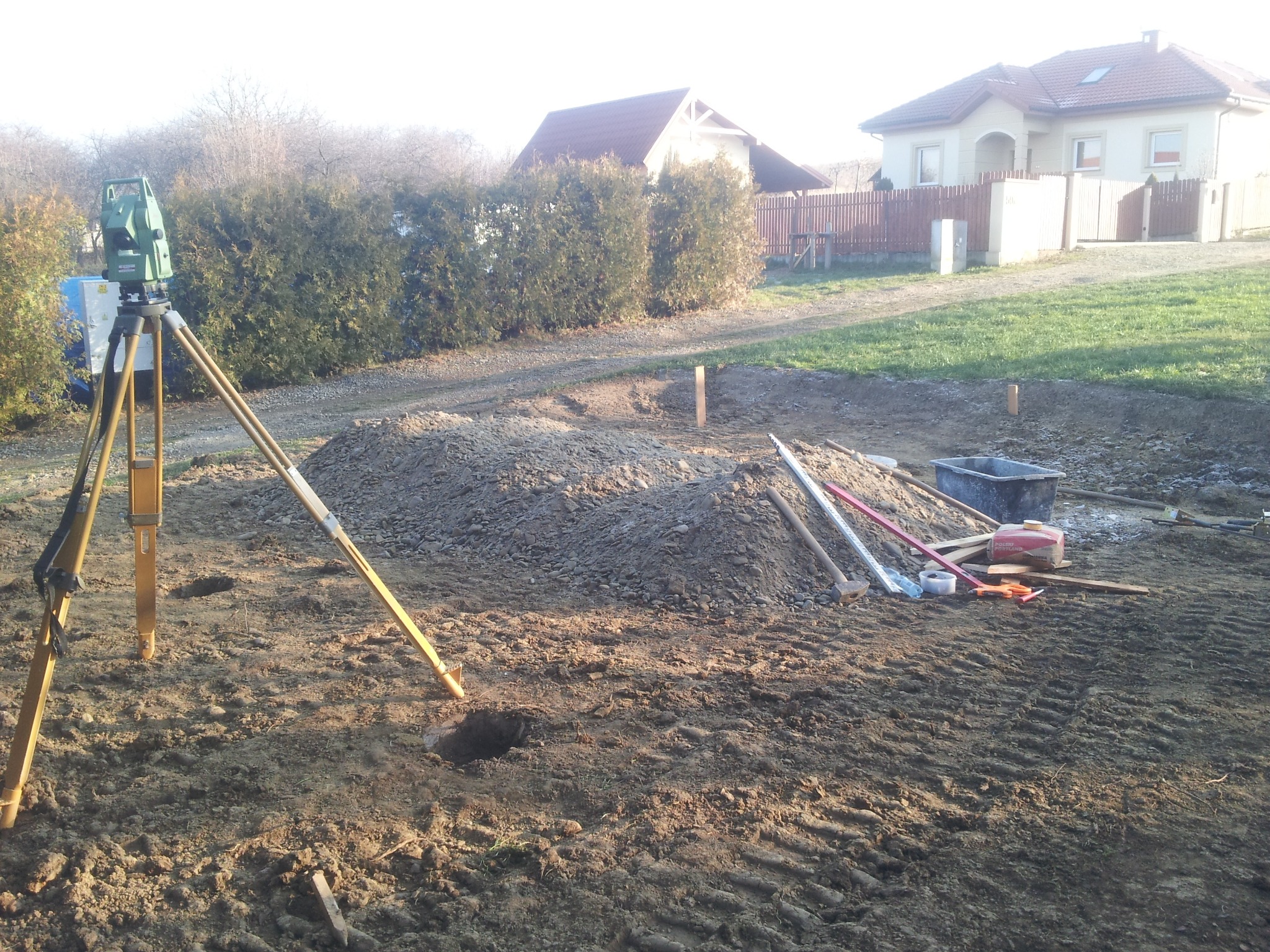 Niwelator optyczny na statywie wyznacza punkty na budowie domu jednorodzinnego z czerwonym dachem, widoczne narzędzia i wykop