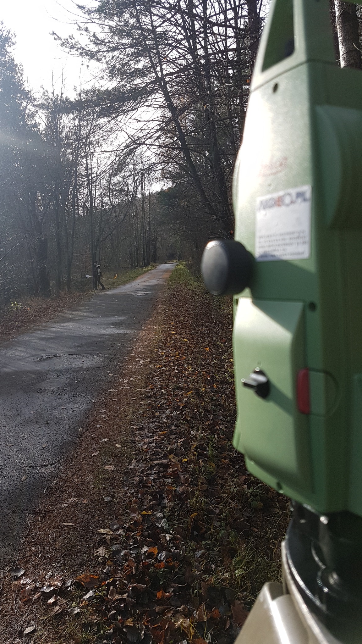 Zielony teodolit na tle leśnej drogi z pracującym geodetą w oddali, jesienne liście na poboczu.