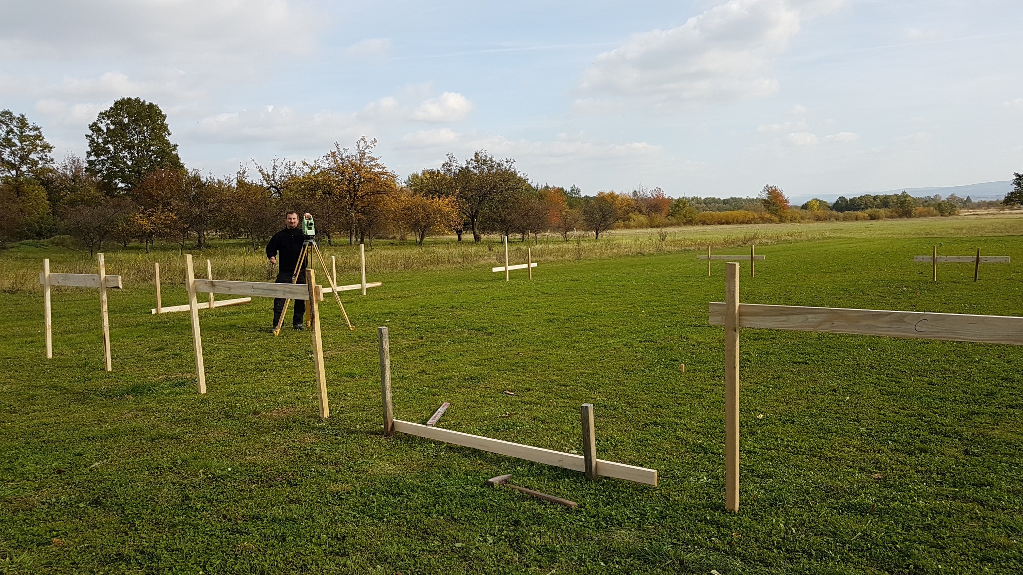 Mężczyzna w czarnym ubraniu używa niwelatora na trójnogu na zielonym polu z drewnianymi palikami, wyznaczając teren pod budowę w słoneczny dzień.