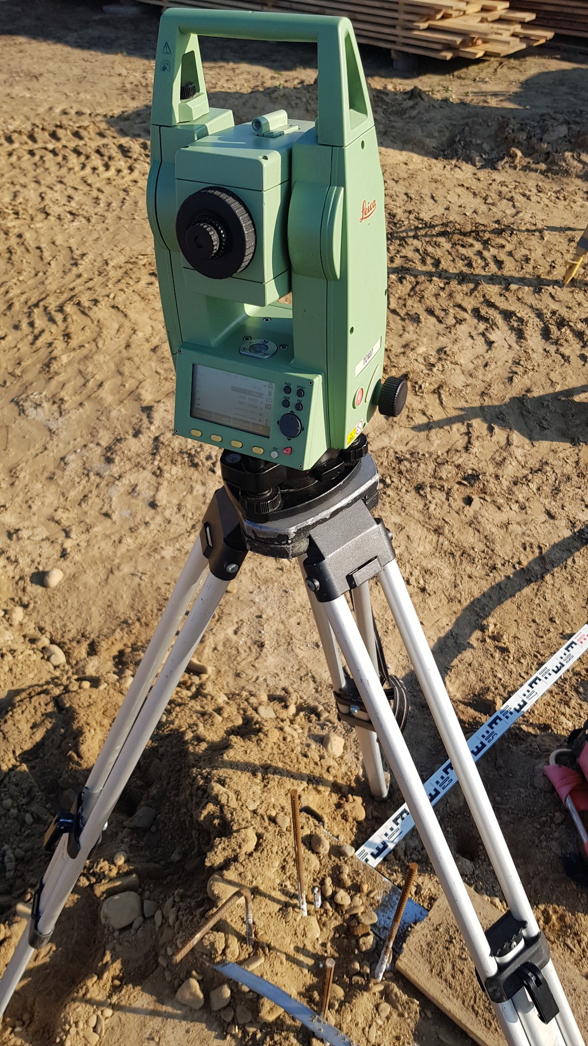 Zielony teodolit Leica TC407 na trójnogu, ustawiony na piaszczystym terenie budowy, obok leży składana łata geodezyjna i pręty zbrojeniowe.