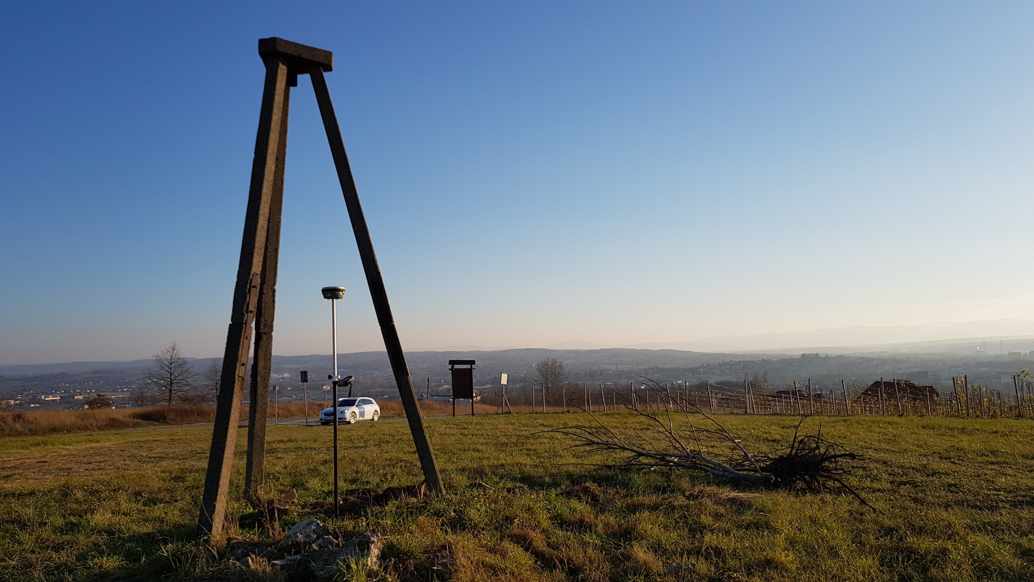 Statyw geodezyjny z odbiornikiem GNSS na tle pagórkowatego krajobrazu i białego samochodu, widoczne pozostałości po ściętych gałęziach.