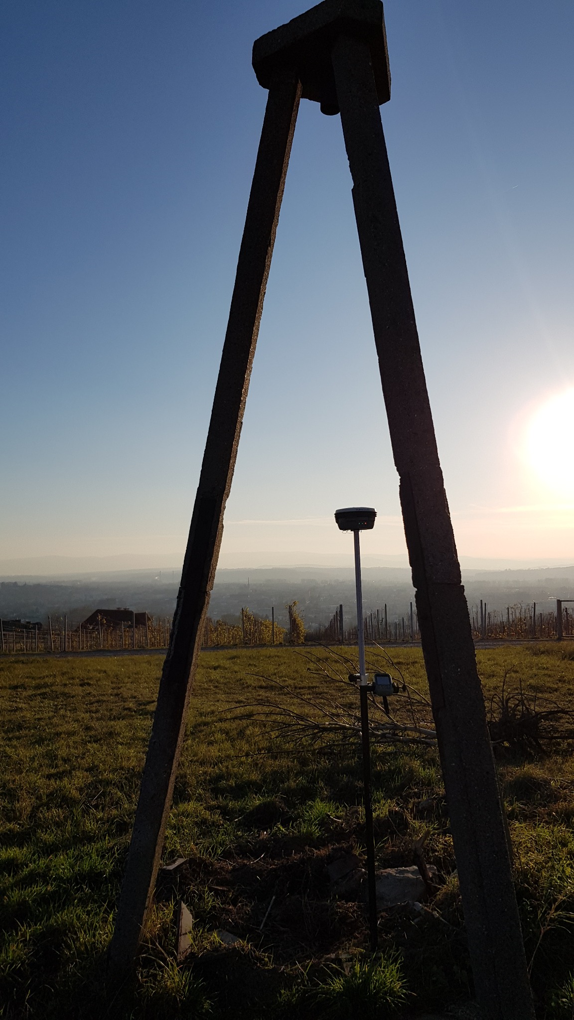 Statyw geodezyjny z odbiornikiem GPS na tle winnicy i odległych zabudowań miejskich w promieniach zachodzącego słońca.