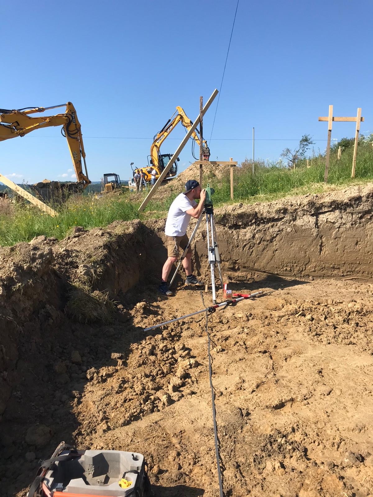 Pracownik w białej koszulce i beżowych szortach obsługuje niwelator na statywie, w tle wykop i dwie koparki JCB na placu budowy.