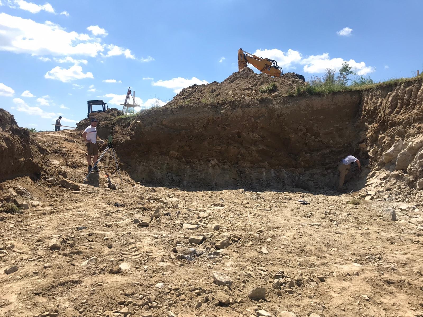 Prace geodezyjne w Jasło: geodeta z niwelatorem na tle wykopu, w tle koparka i inni pracownicy.