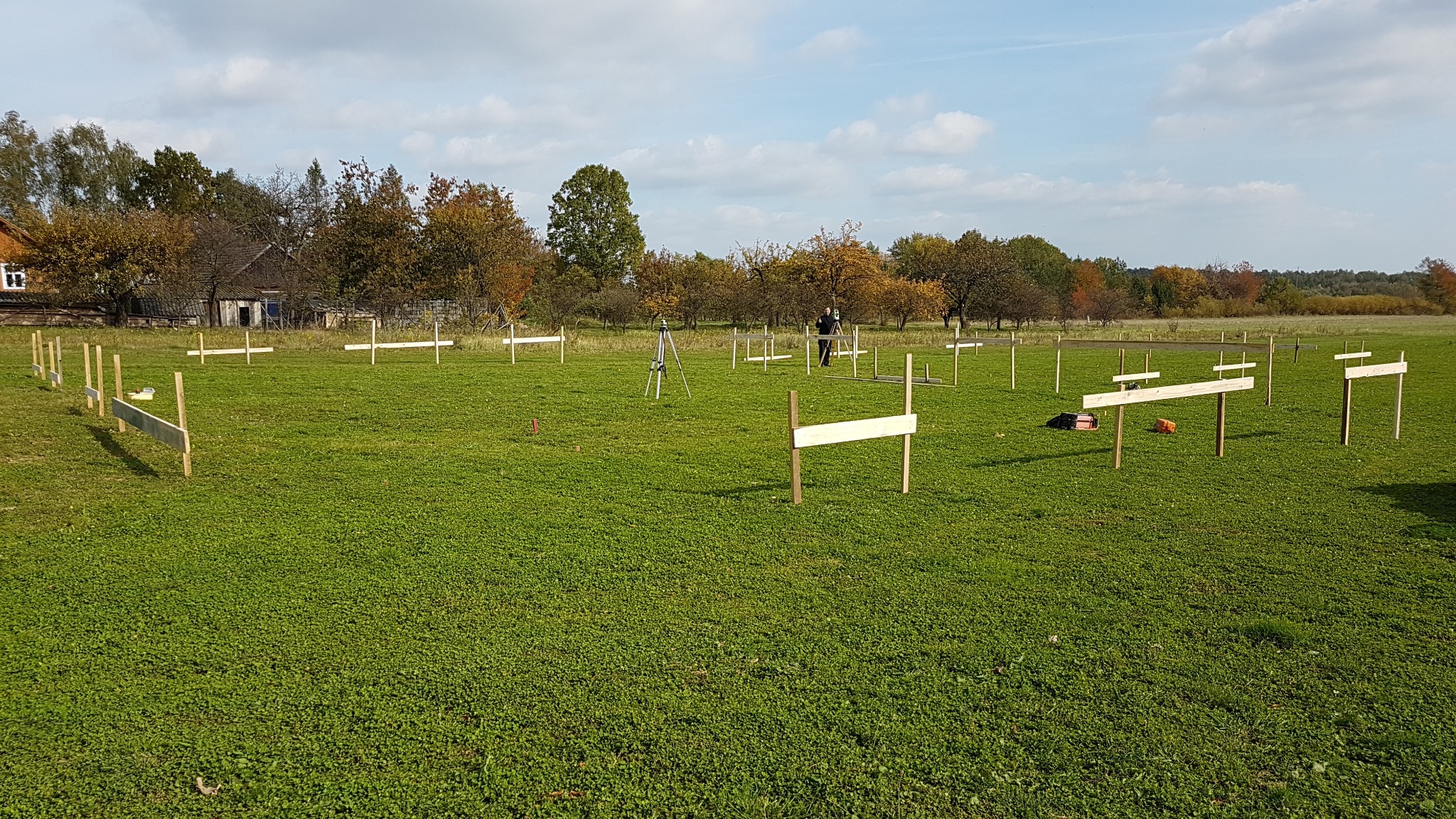 Wytyczanie granic działki: drewniane znaczniki wbite w trawiastą łąkę, w tle drzewa i budynek, widoczny teodolit na statywie oraz osoba obsługująca sprzęt pomiarowy.