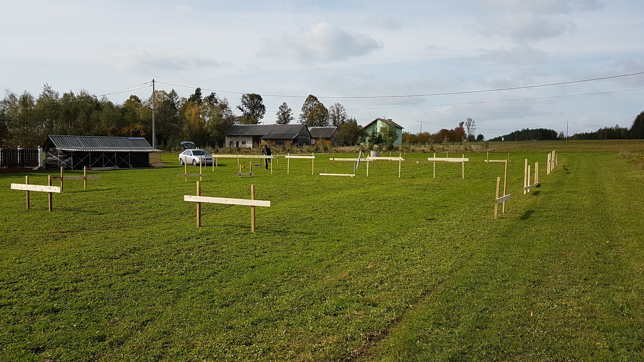 Wytyczanie fundamentów budynku na zielonej łące, widoczne drewniane pale i poziome deski, w tle zabudowania gospodarcze i samochód z otwartym bagażnikiem.