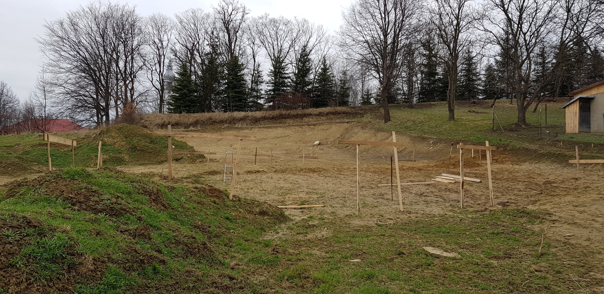 Wytyczanie fundamentów budynku na działce, widoczne drewniane słupki i poprzeczki, w tle kościół i drzewa.