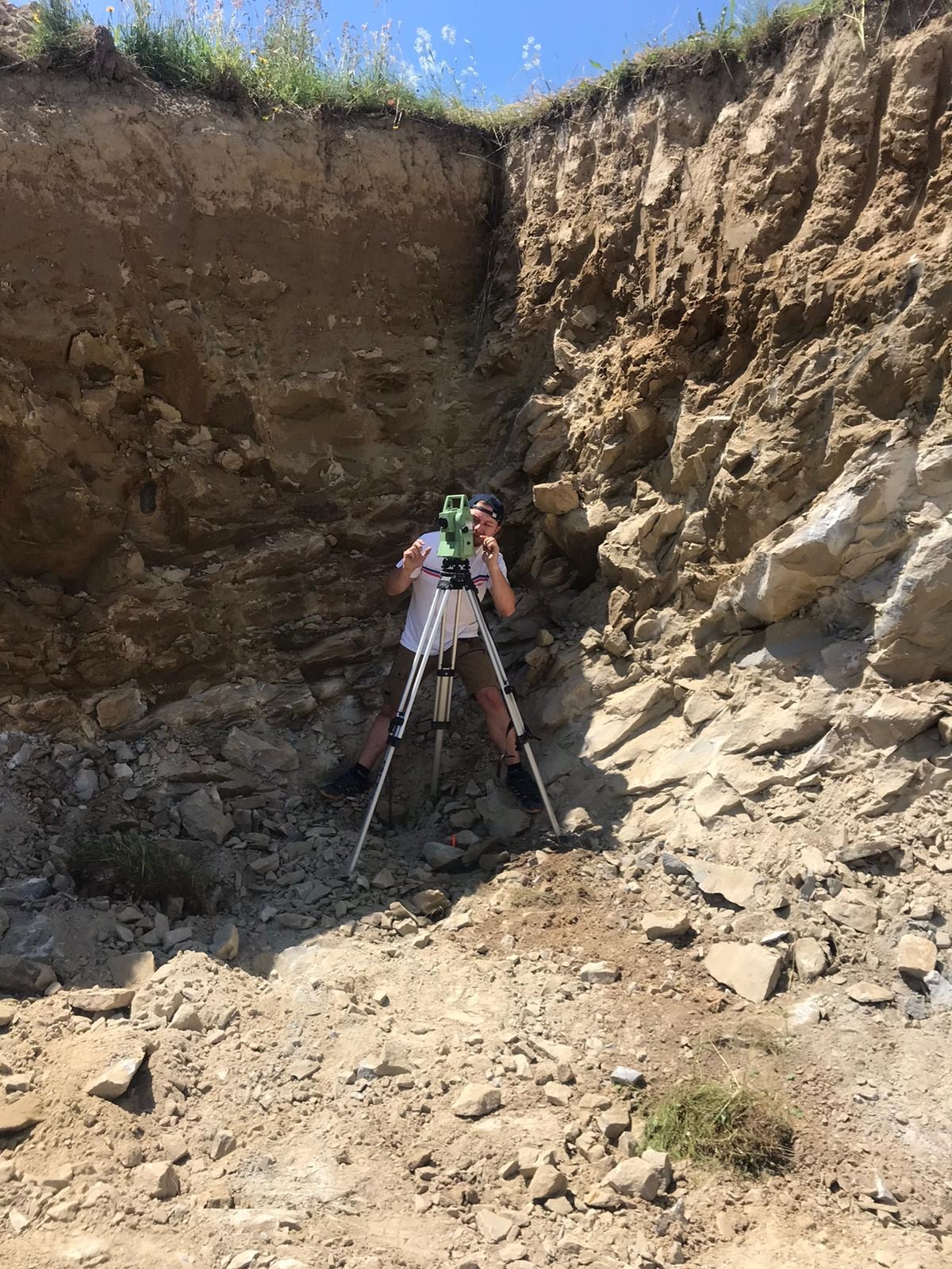 Mężczyzna w krótkich spodenkach i koszulce z krótkim rękawem obsługuje teodolit na statywie w wykopie o skalistych ścianach.