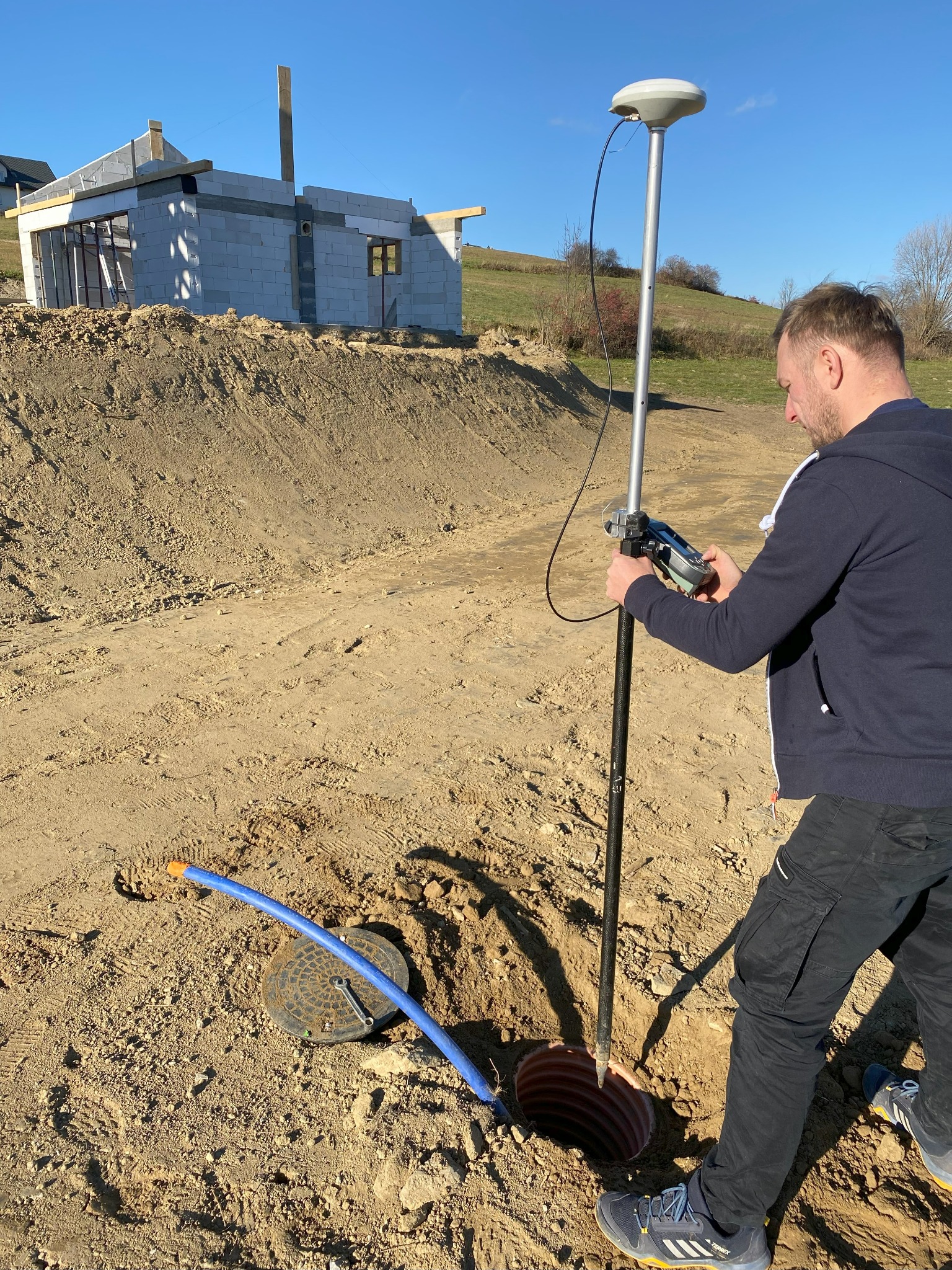 Pracownik w ciemnej bluzie obsługuje precyzyjny instrument pomiarowy nad studzienką kanalizacyjną na placu budowy domu z białego pustaka.