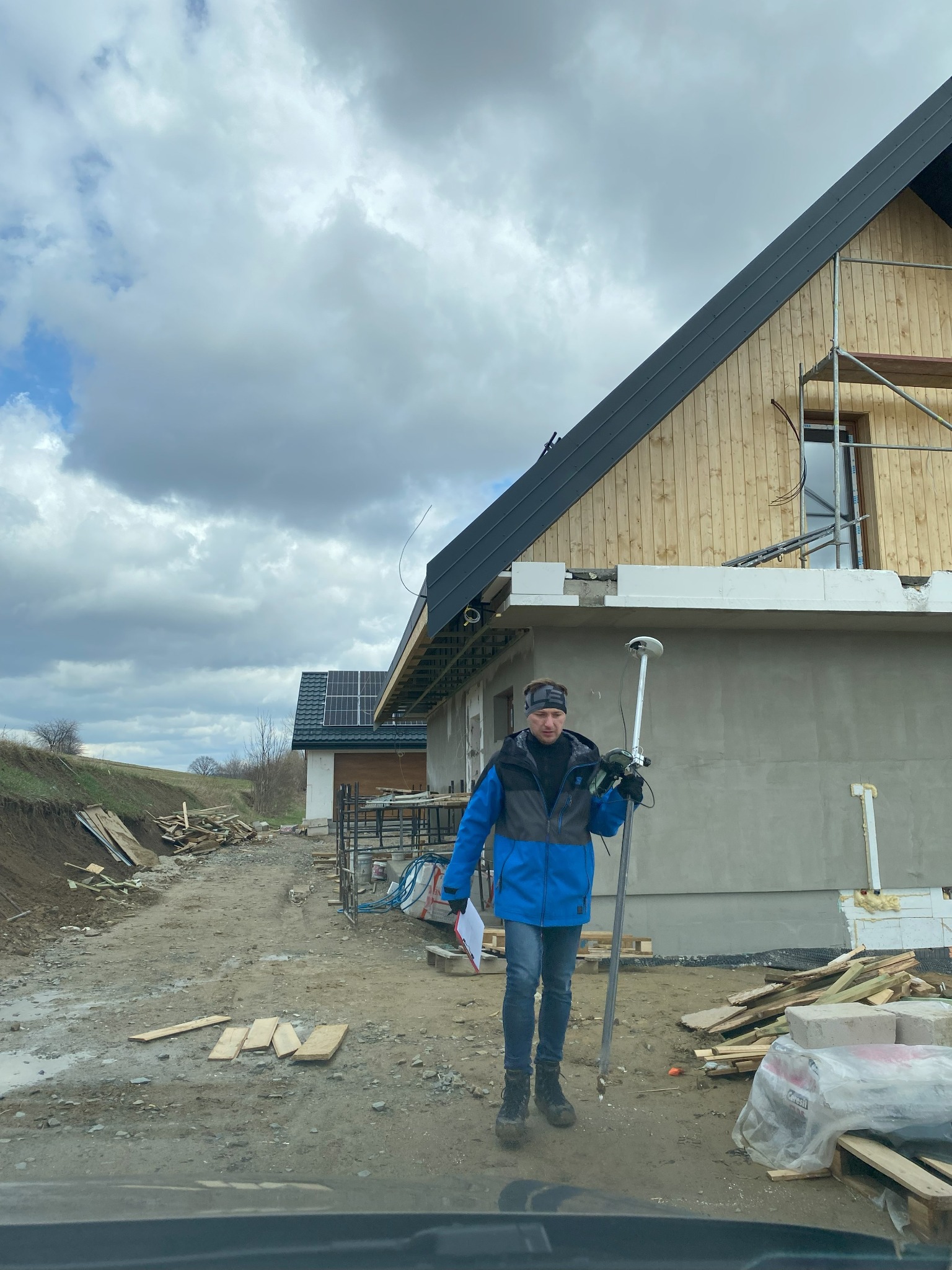 Geodeta w niebieskiej kurtce z niwelatorem na tle surowego stanu domu z drewnianą elewacją i szarym dachem w pochmurny dzień.