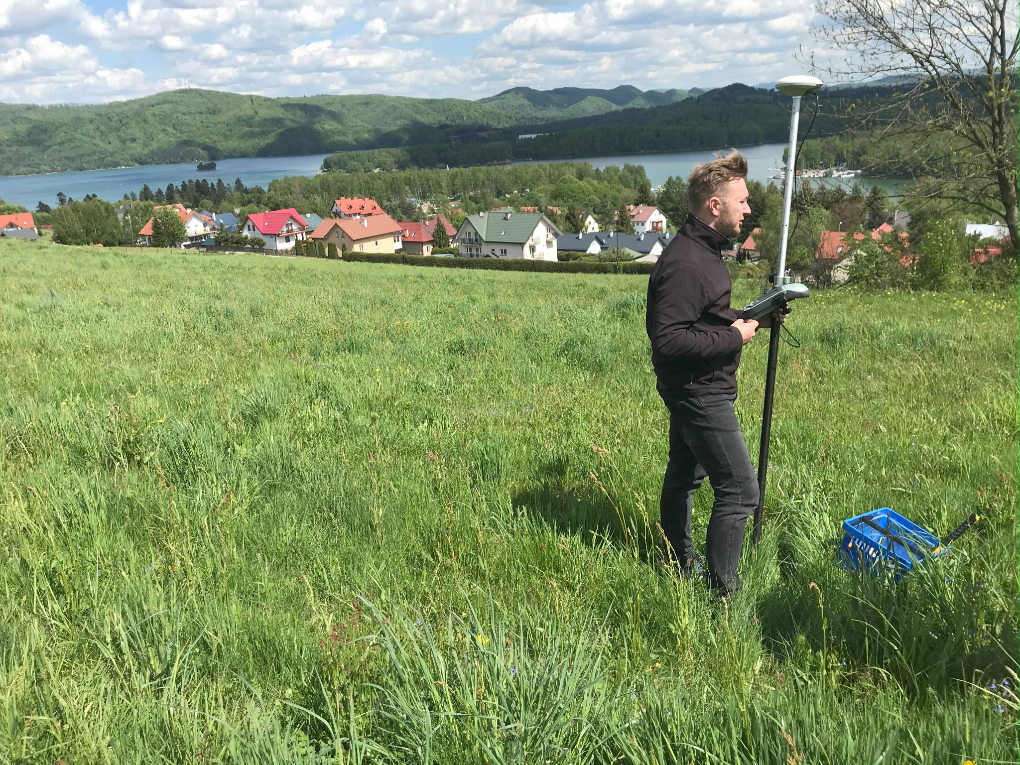 Mężczyzna w czarnej kurtce i jeansach obsługuje sprzęt pomiarowy GPS na łące z widokiem na jezioro i zabudowania w tle.