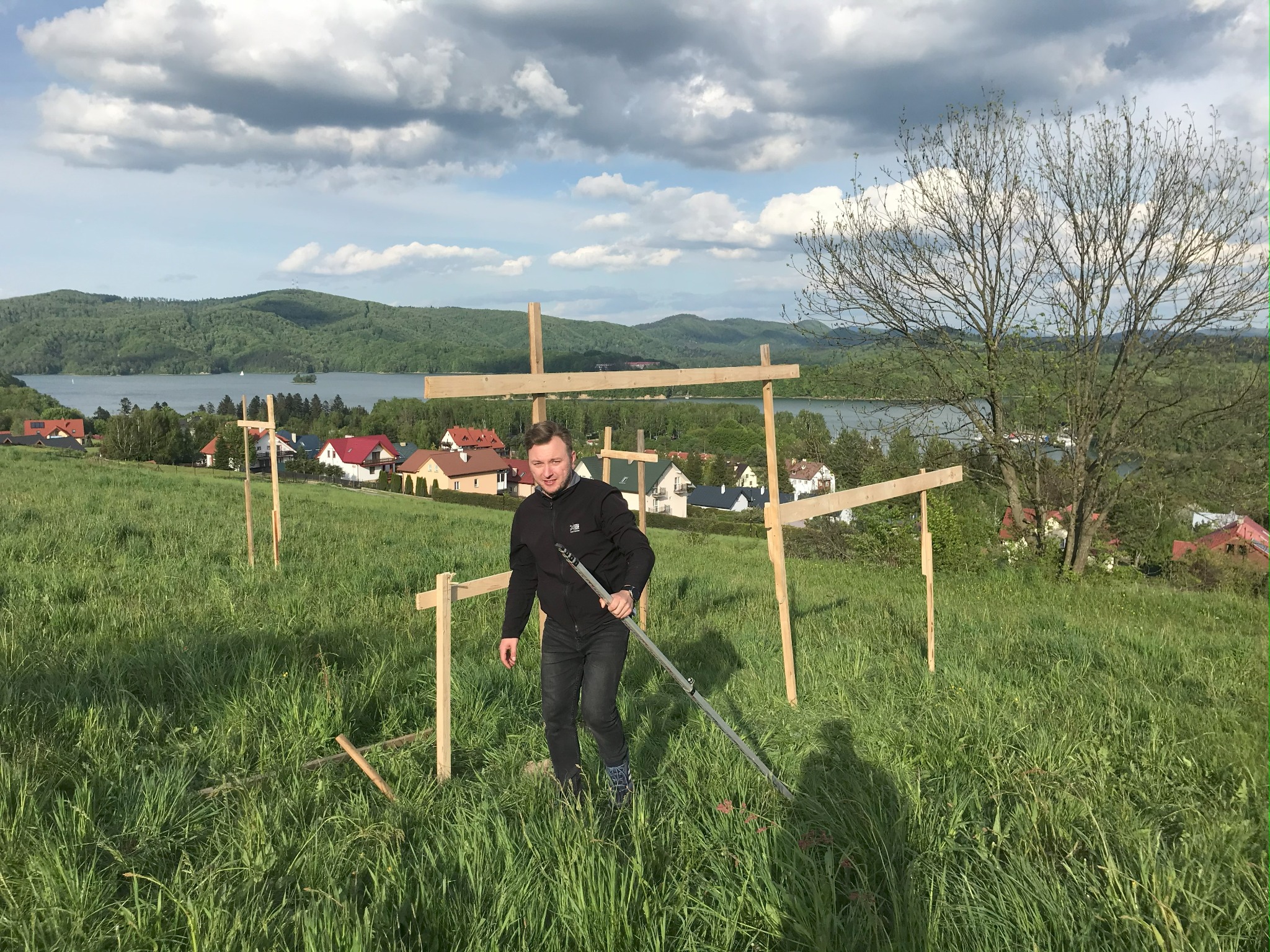 Mężczyzna z niwelatorem tyczkowym na łące z drewnianymi konstrukcjami, w tle jezioro i domy.