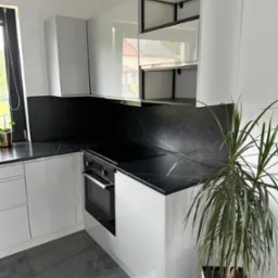 Biała, nowoczesna kuchnia z czarnym blatem imitującym marmur, płyta indukcyjna i piekarnik w zabudowie, szare płytki podłogowe, roślina doniczkowa w białej donicy.