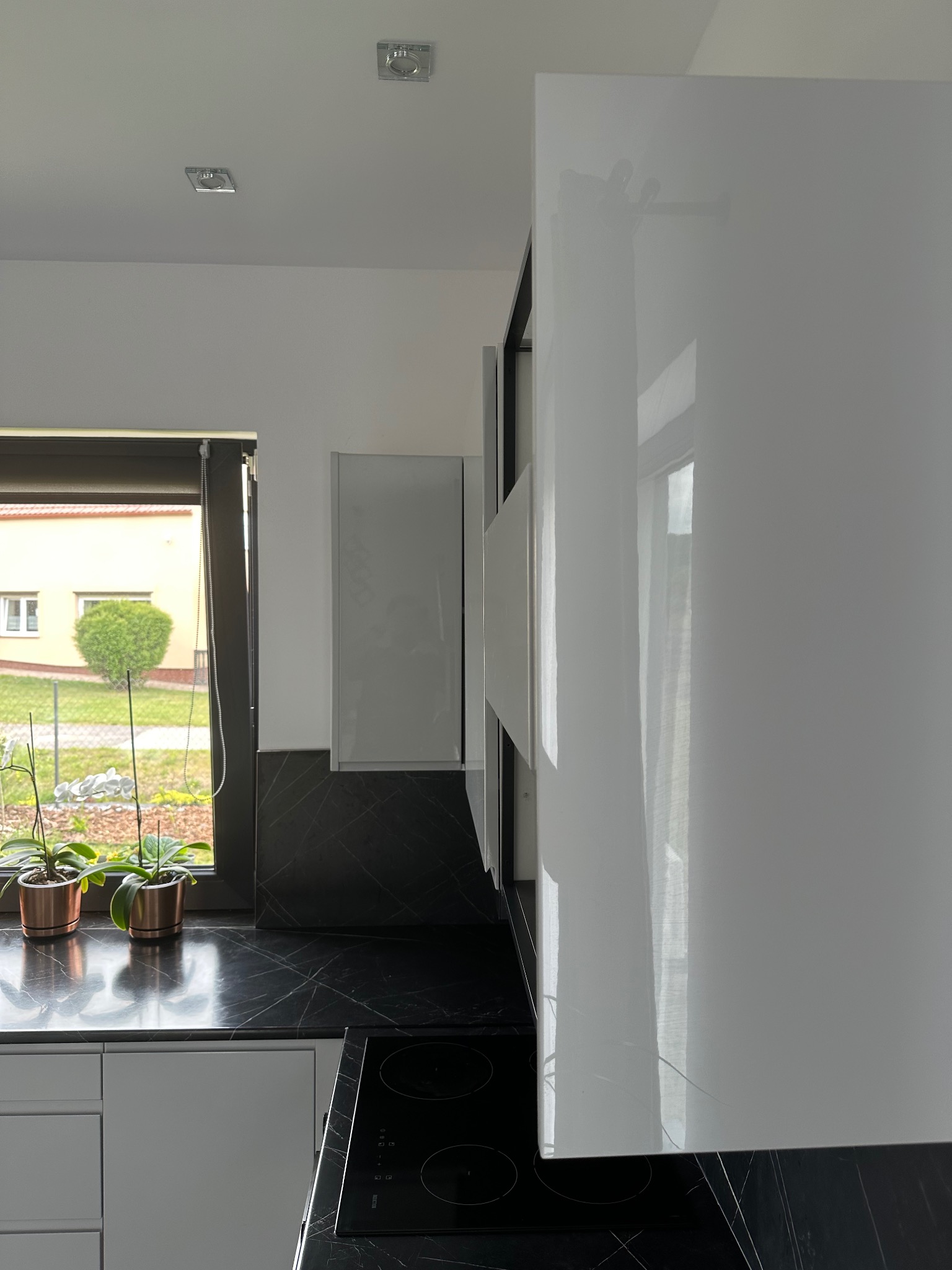Fragment nowoczesnej kuchni z białymi, lakierowanymi szafkami i czarnym blatem imitującym marmur, widok z boku, z oknem i roślinami w tle.