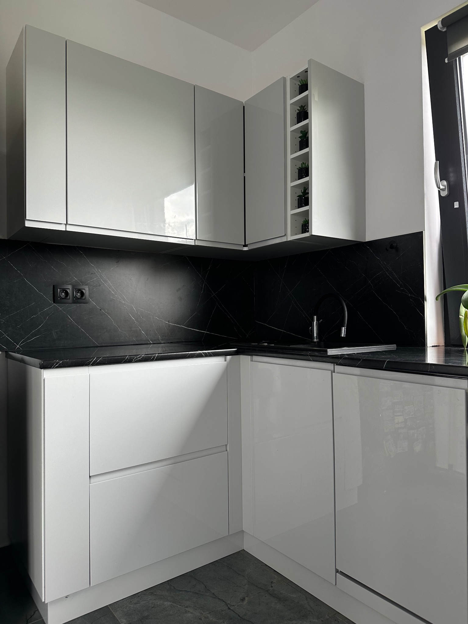 Biała, błyszcząca zabudowa kuchenna narożna z czarnym blatem imitującym marmur, z otwartą szafką na zioła w doniczkach i czarną armaturą.