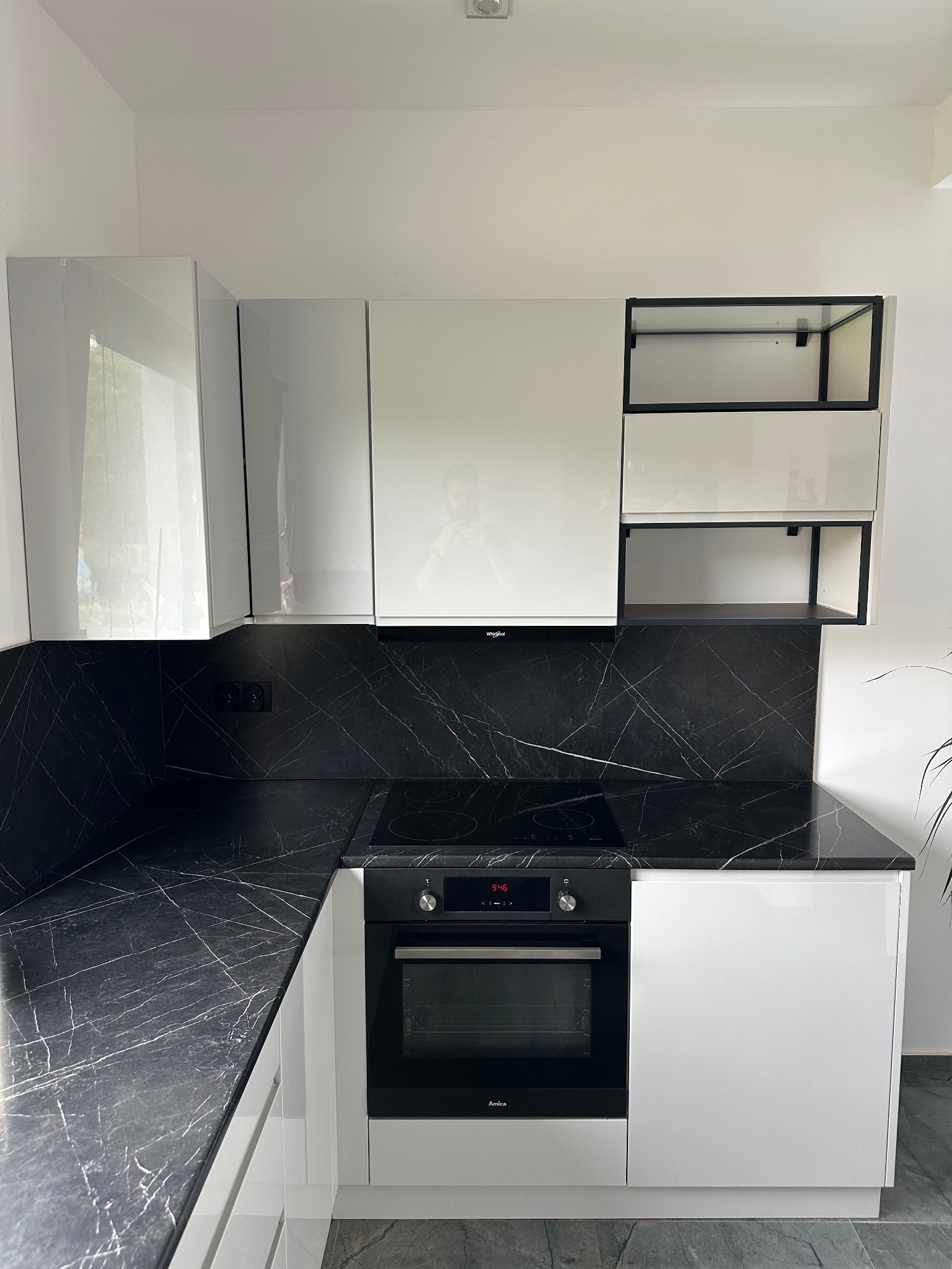 Białe, lakierowane szafki kuchenne z czarnym blatem i backsplashiem imitującym marmur, z piekarnikiem Amica w zabudowie.